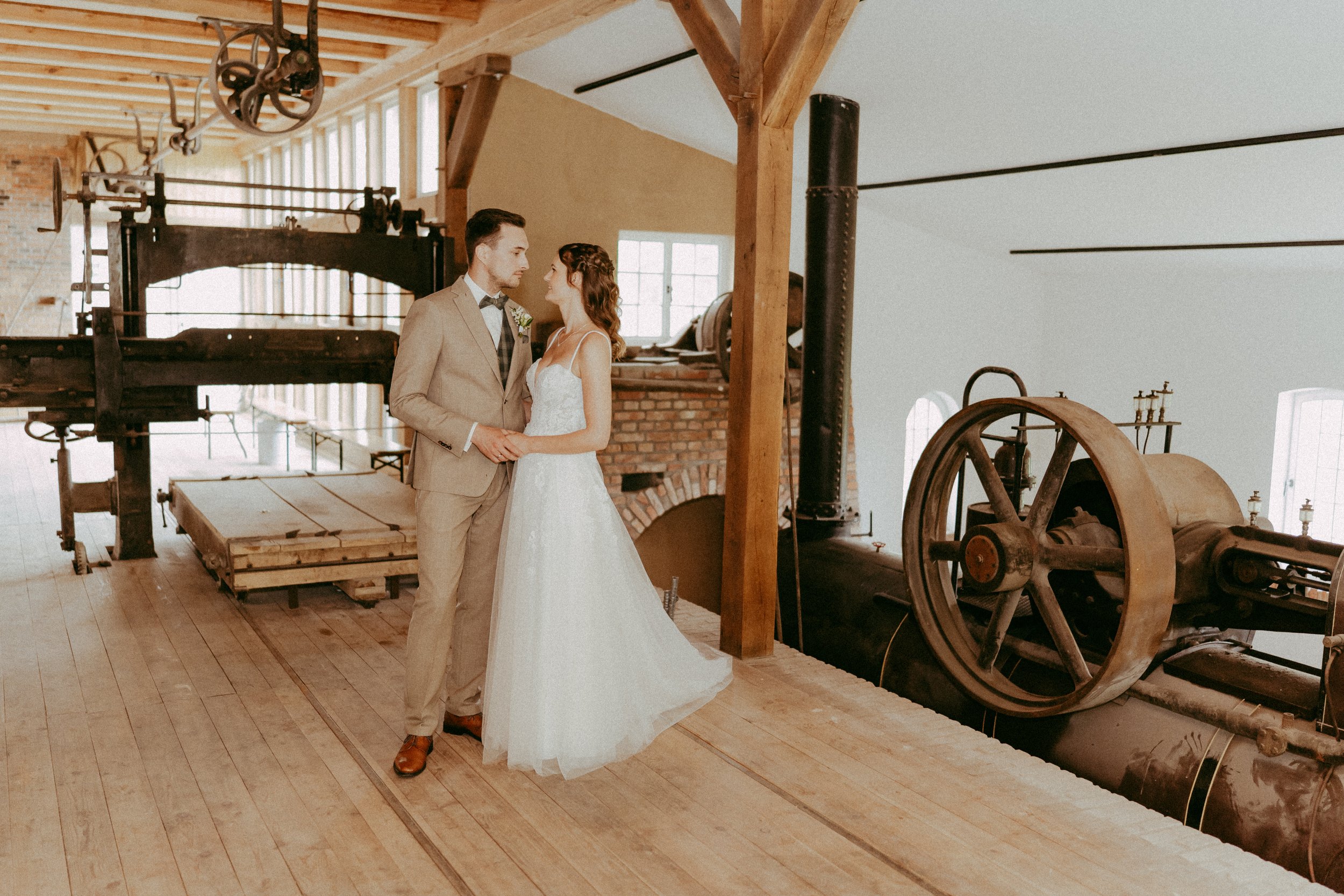 Ein Brautpaar in Hochzeitskleidung steht in einem alten industriellen Gebäude neben einer historischen Maschine.