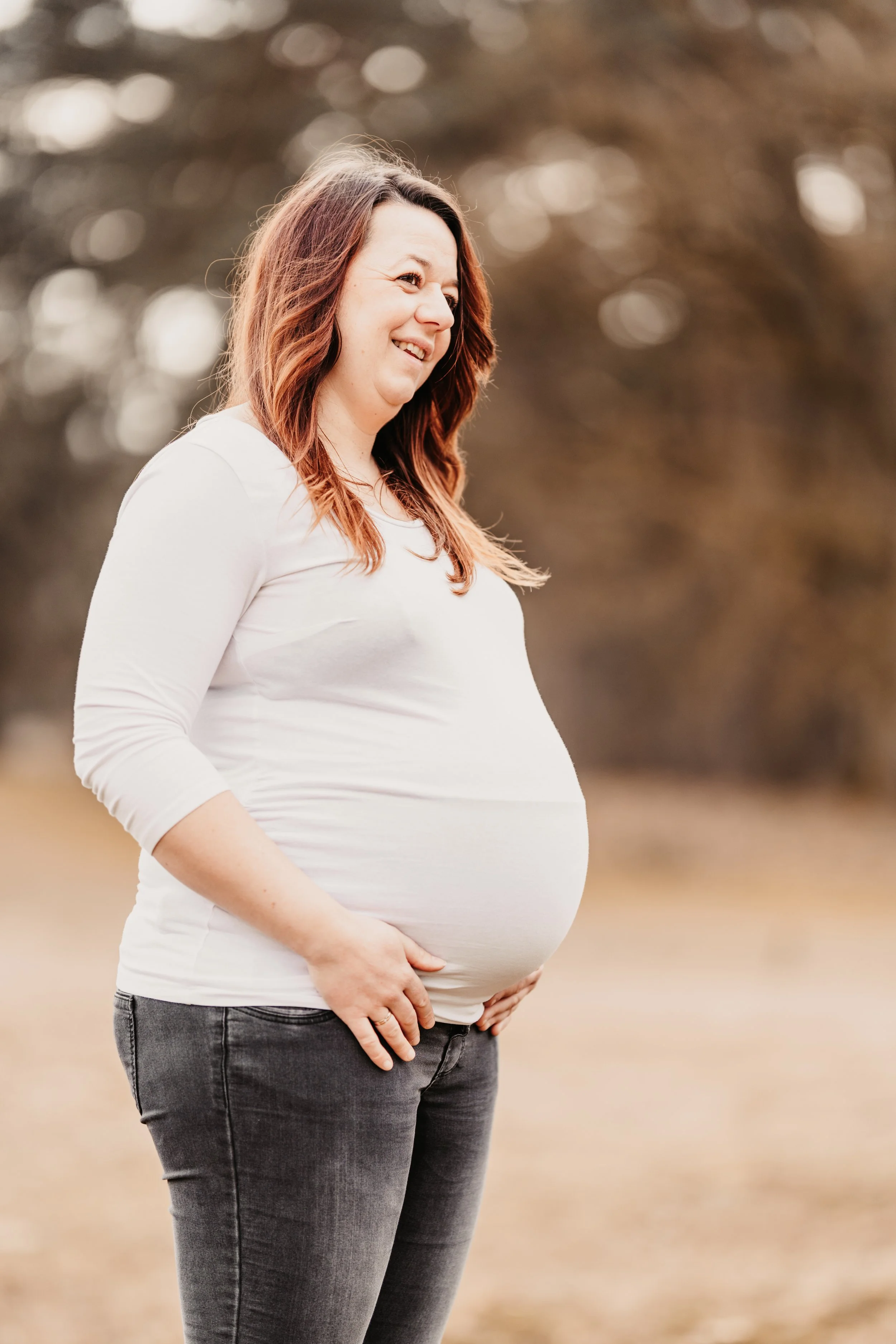 Schwangere Frau mit braunen Haaren in einem weißen Langarmshirt und Jeans steht im Freien, lächelt und schaut zur Seite.
