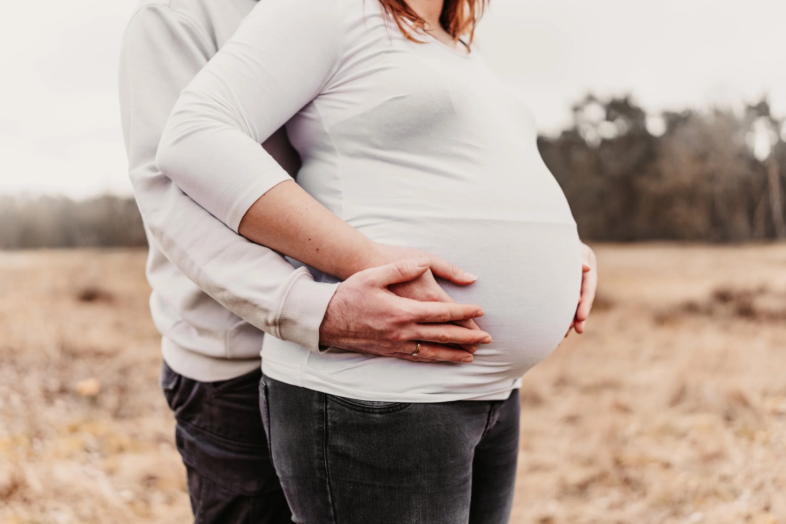 Ein Paar hält bei Sonnenuntergang den Bauch einer schwangeren Frau in einem offenen Feld voller trockener Pflanzen