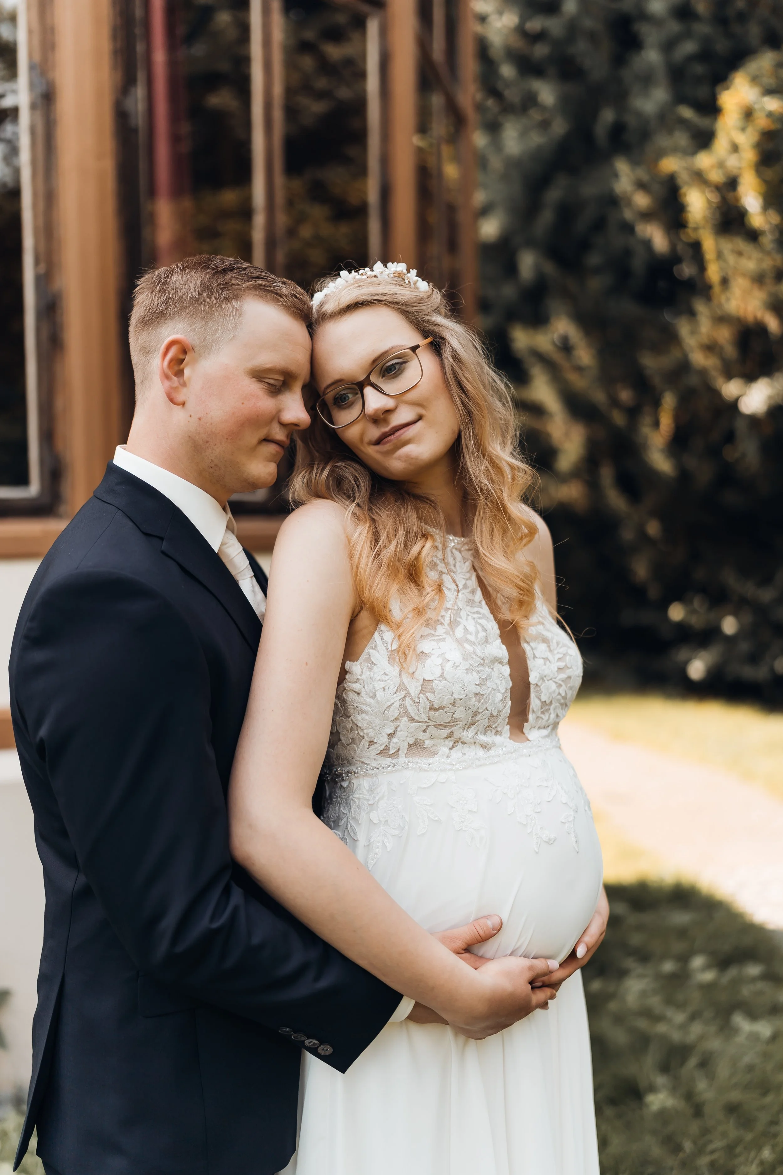 Ein verlobtes Paar, der Mann trägt einen schwarzen Anzug, die Frau ein weißes Hochzeitskleid, umarmt sich vor einem Haus mit offenen Fenstern, sie hält ihren Babybauch. Sie stehen eng beieinander, mit geschlossenen Augen, im Freien bei Tageslicht.