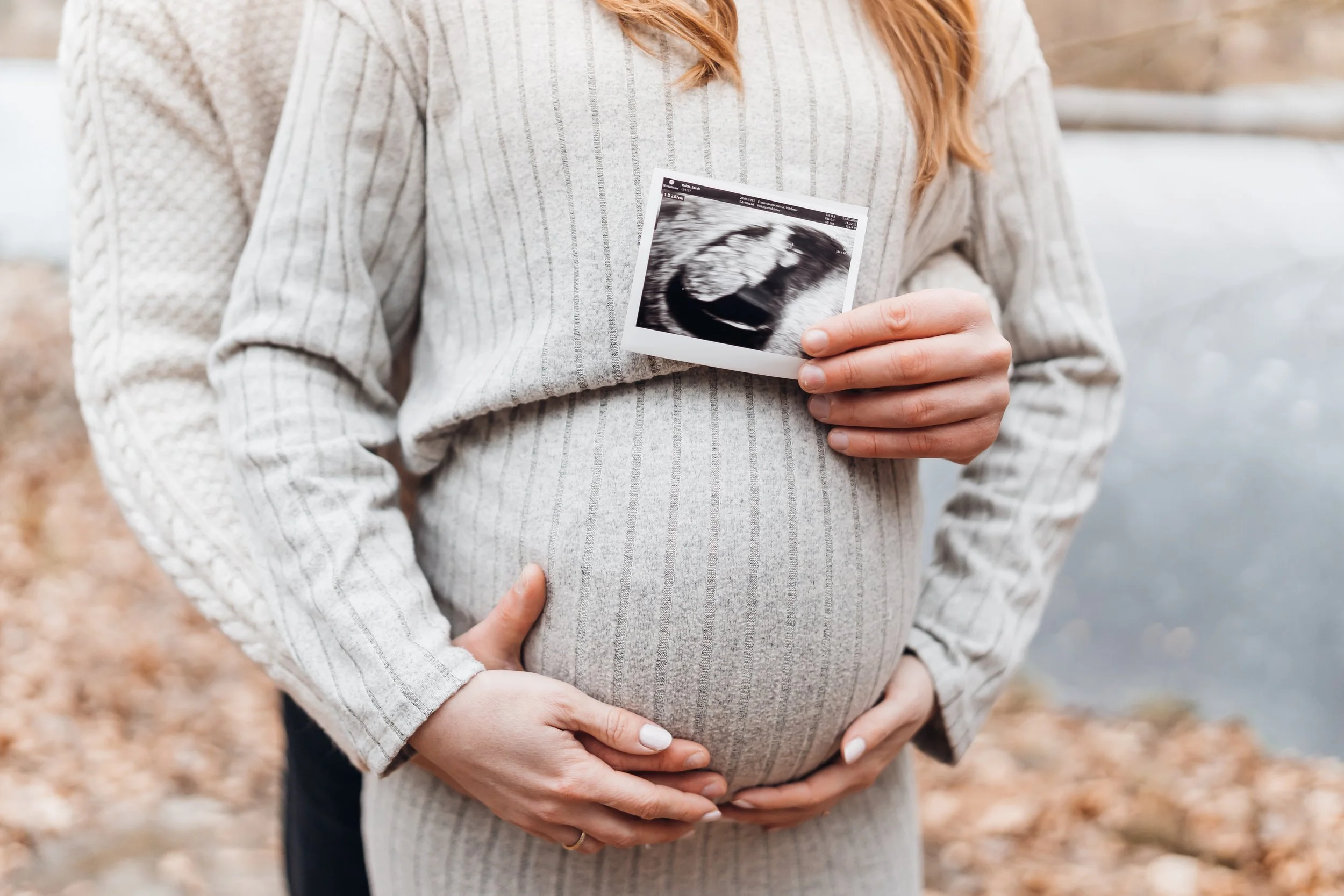 Eine schwangere Frau hält ein Ultraschallbild ihres ungeborenen Kindes vor ihrem Bauch, während sie in der Natur steht.