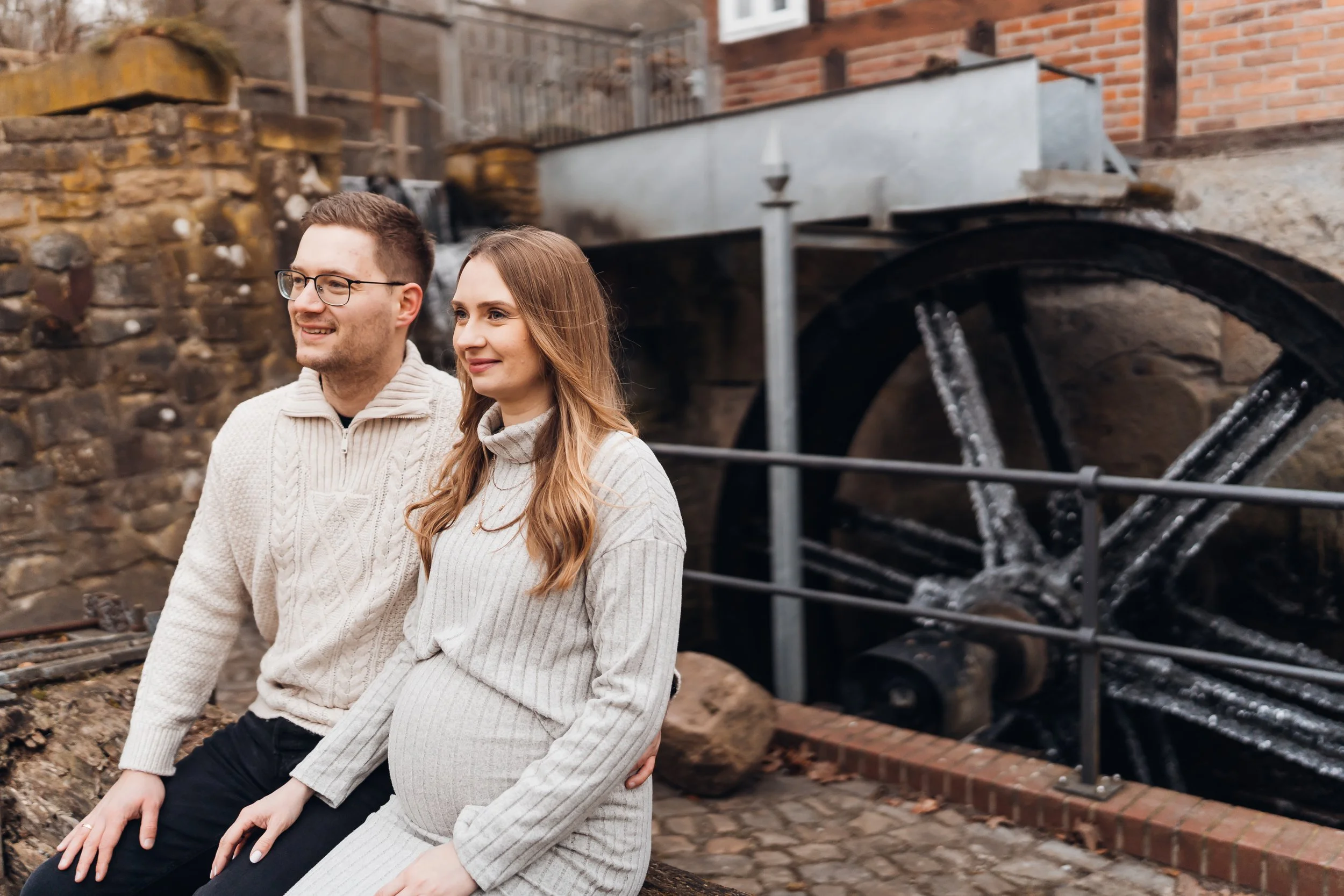 Ein schwangeres Paar sitzt vor einer alten Wasser- oder Mühlrad an einer Steinmauer, im Hintergrund ein Wasserlauf und Gebäudestrukturen.