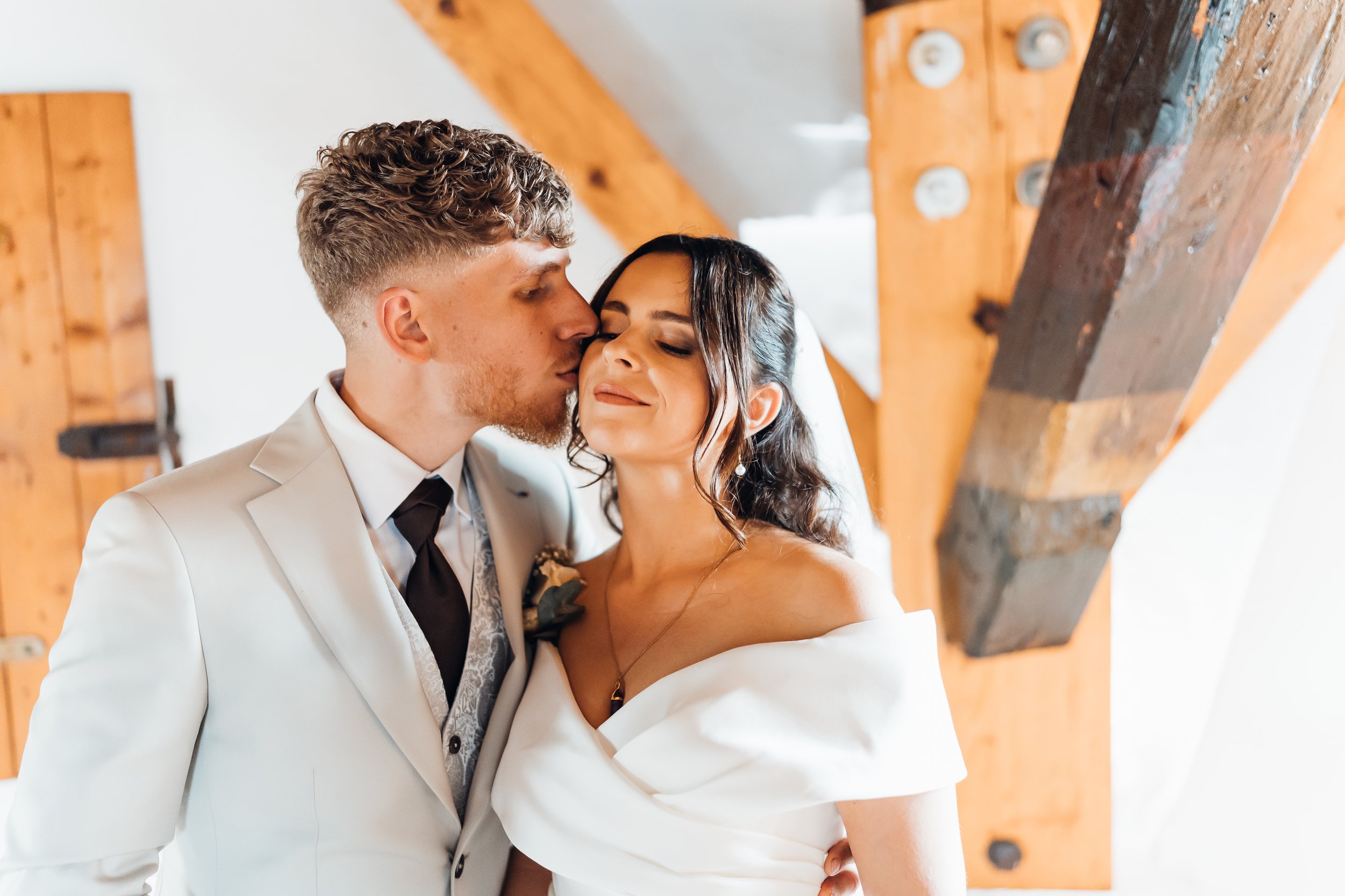 Ein Brautpaar auf einer Hochzeit, der Bräutigam küsst die Braut auf die Stirn, im Hintergrund Holzwände.