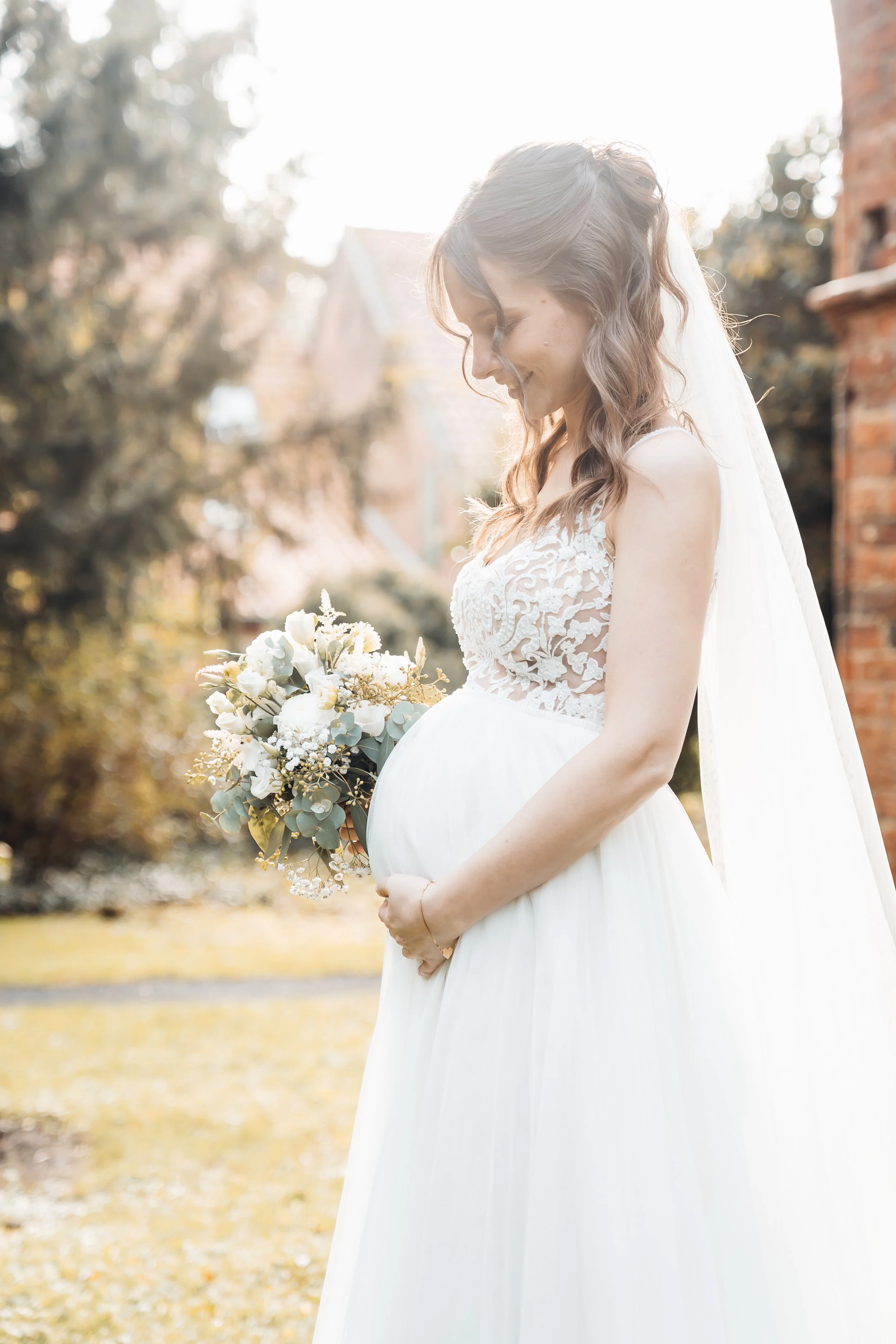 Eine hochschwangere Frau in einem weißen Brautkleid hält einen Blumenstrauß und steht im Sonnenschein vor einem Gebäude mit alten Ziegelwänden im Freien.