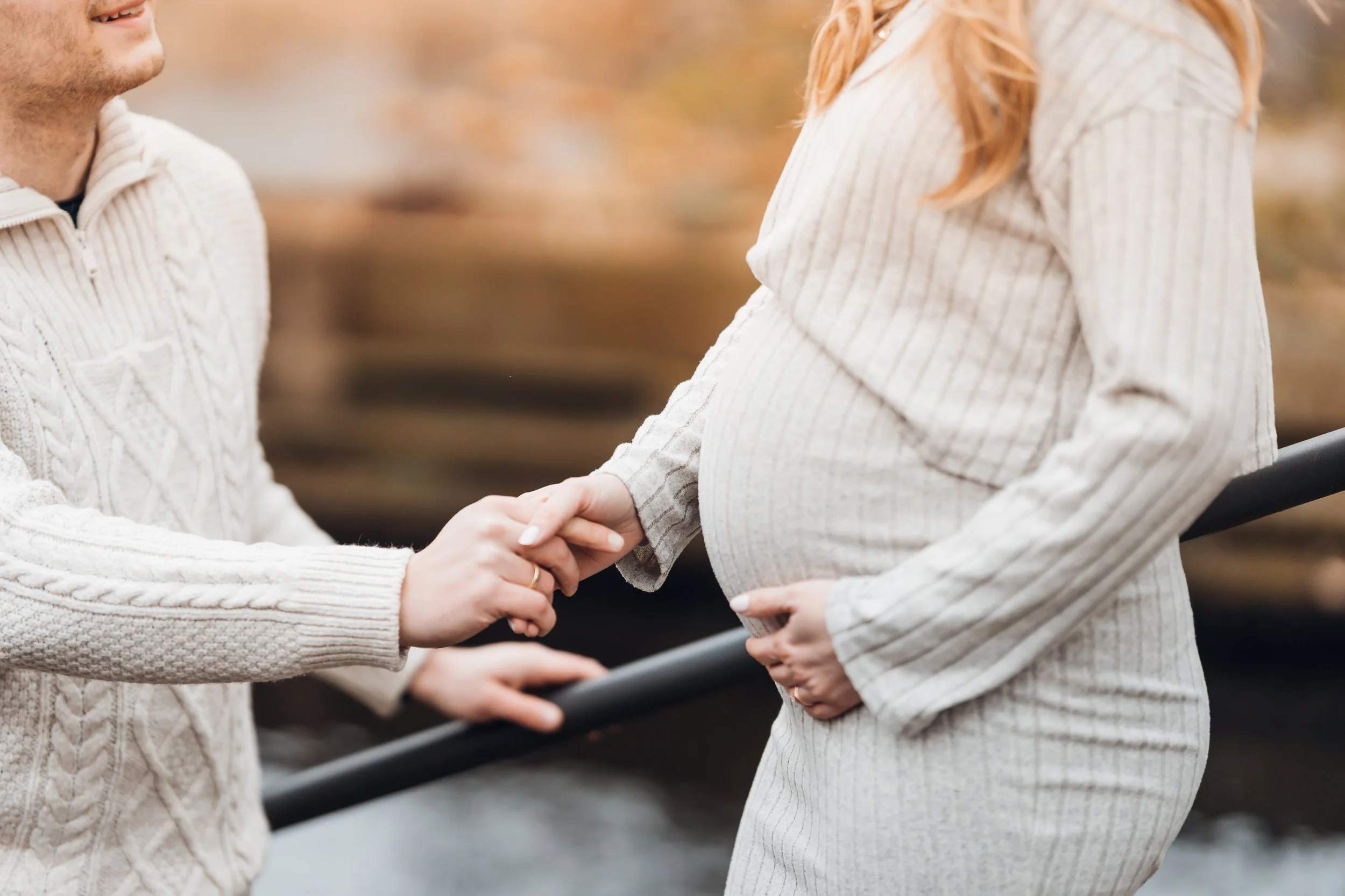 Eine schwangere Frau und ein Mann halten sich an einer Brüstung am Flussufer. Der Mann hält die Hand der Frau.