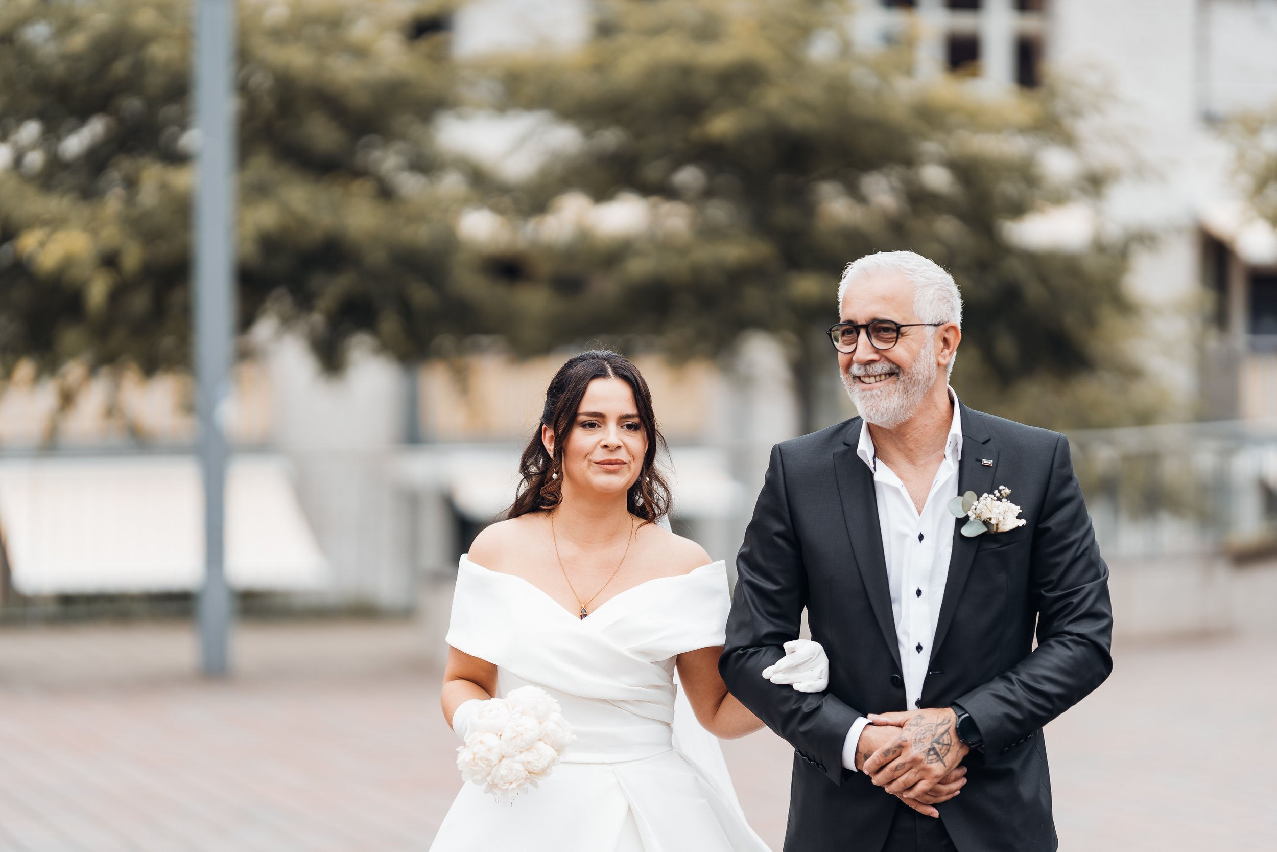 Braut und älterer Mann bei einer Hochzeit, die Frau trägt ein weißen Hochzeitskleid und hält einen Blumenstrauß, der Mann trägt einen schwarzen Anzug mit weißem Hemd, beide lächeln und sind draußen bei einer Hochzeit.