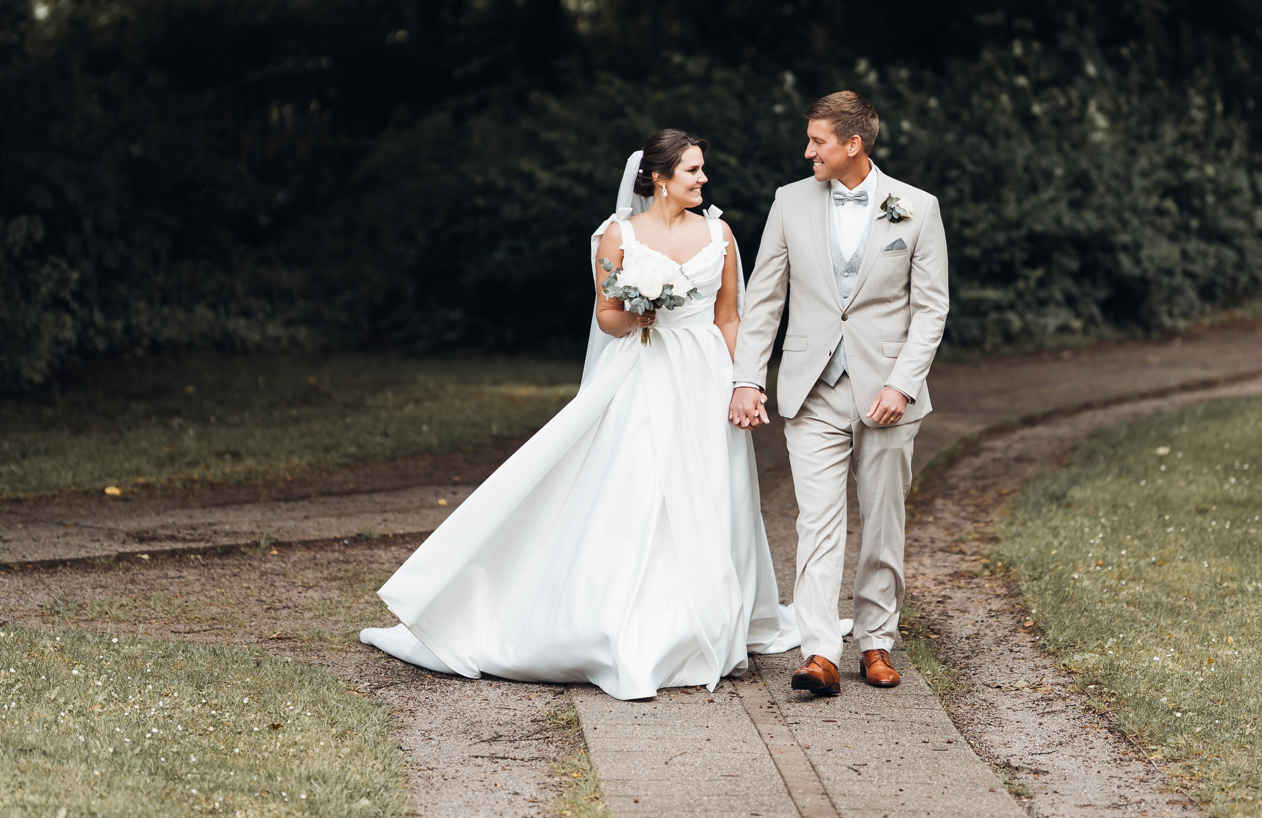 Ein junges Brautpaar, das Hand in Hand auf einem Weg in einem Park spaziert, lachend und glücklich an ihrem Hochzeitstag.