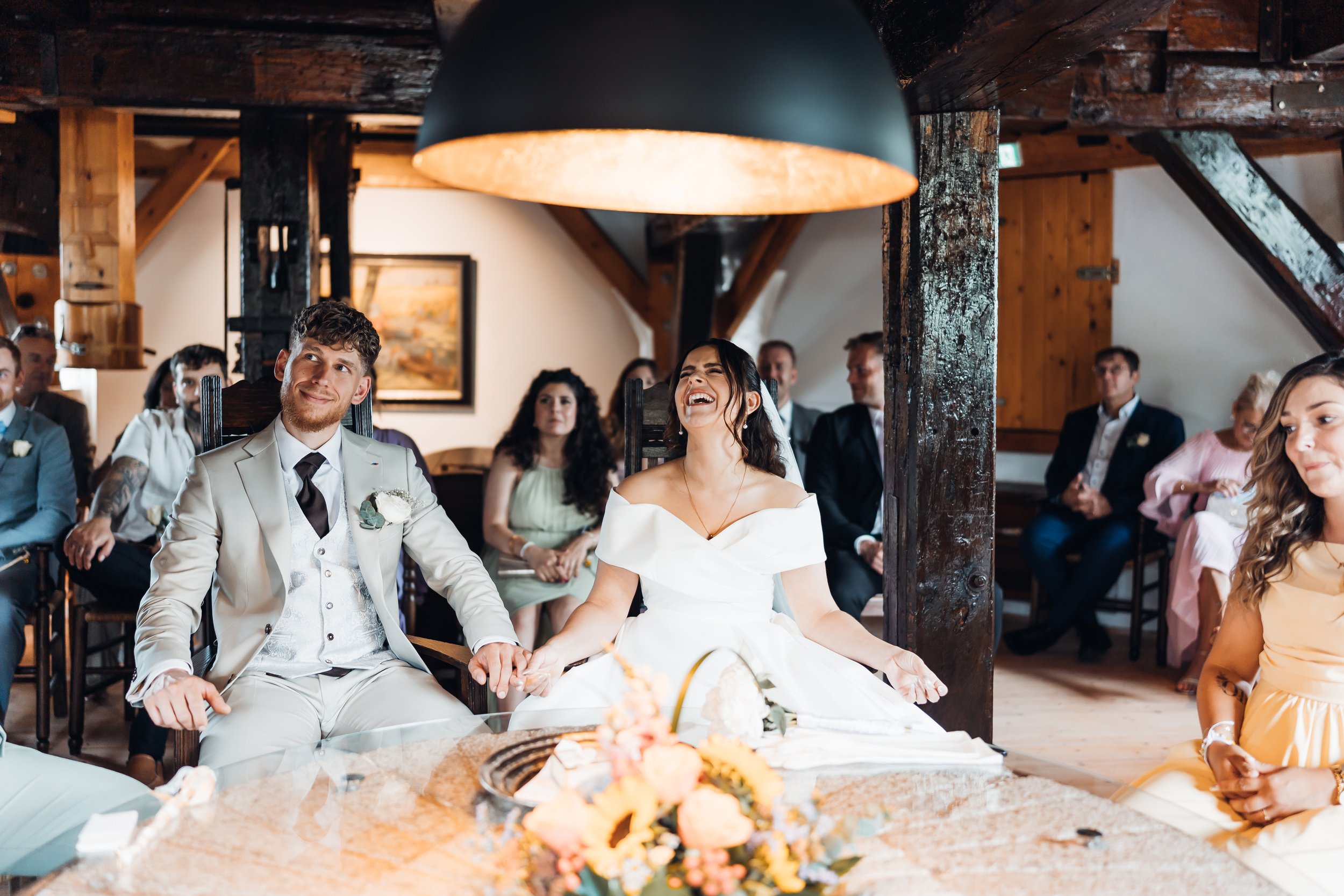 Hochzeitspaar sitzt lachend bei ihrer Trauung in einem rustikalen Raum, umgeben von Gästen.