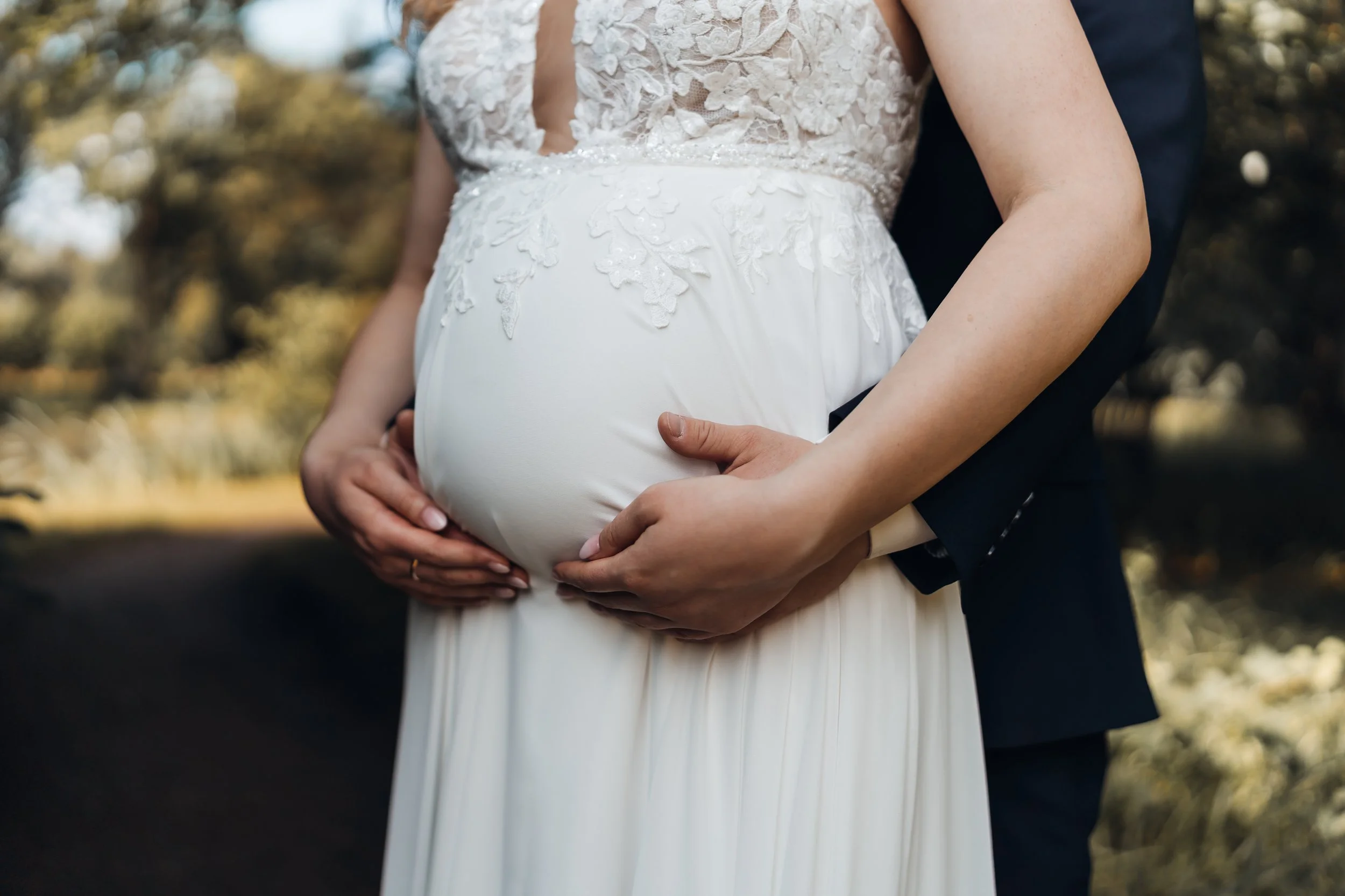 Braut in weißem Kleid hält Babybauch, während ein Mann sie von hinten umarmt in einem Park bei Sonnenuntergang.