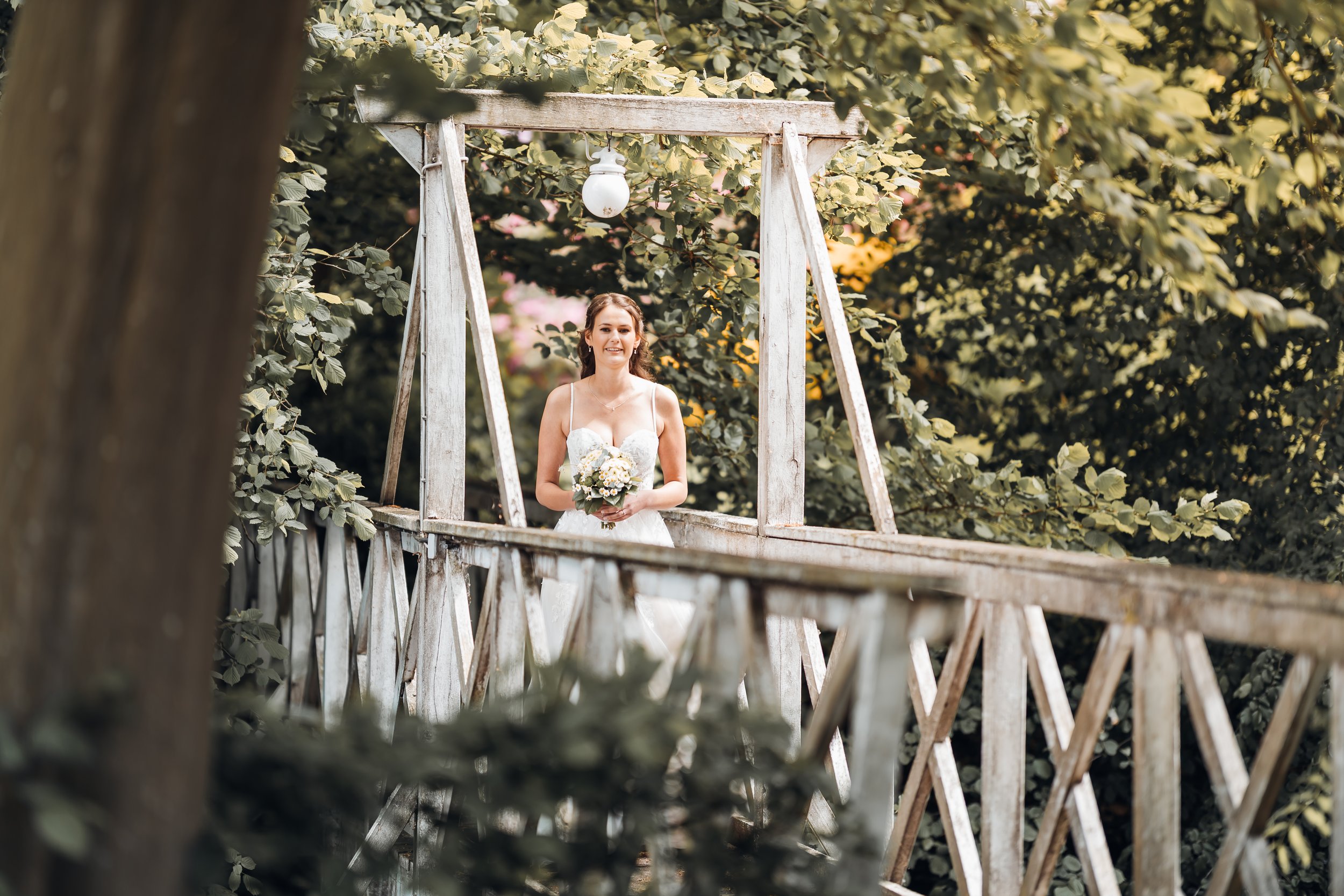 Braut in Hochzeitskleid hält Blumenstrauß auf einer Holzbrücke im Garten, umgeben von Bäumen