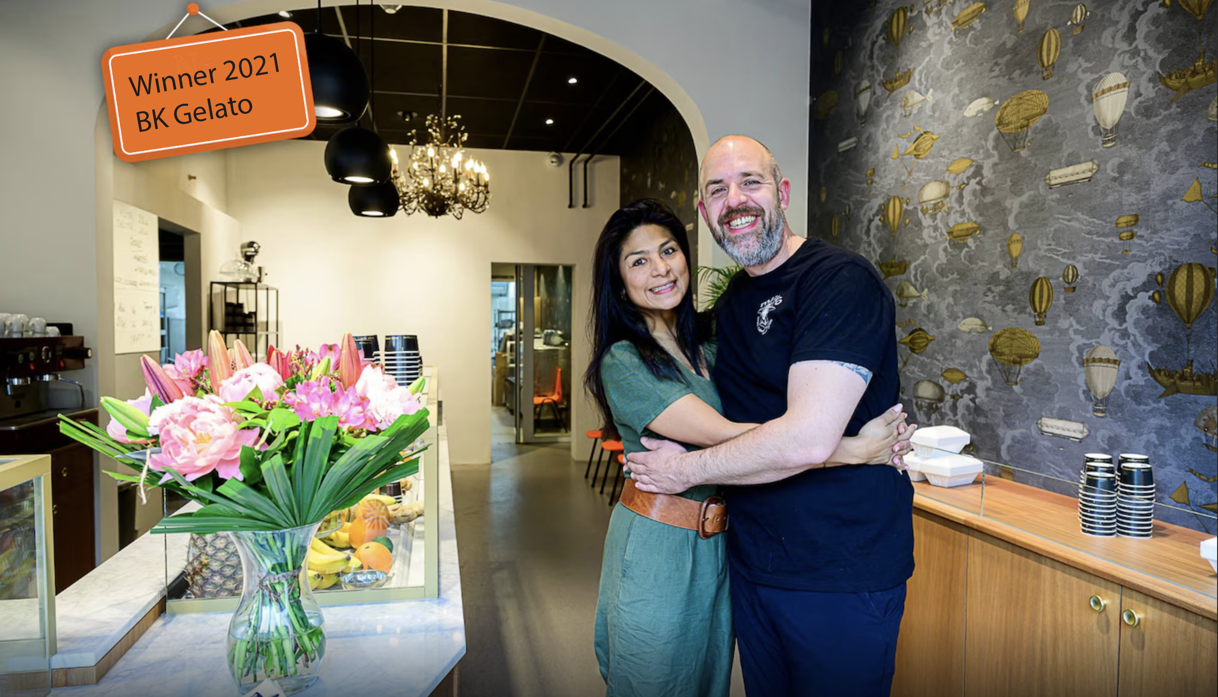 A couple hugging and smiling inside a bakery or cafe. The woman has long dark hair and wears a green dress, and the man has a beard, shaved head, and is wearing a black T-shirt. A large bouquet of pink and white flowers is on the counter in front of them, and there is a sign indicating they are the 2021 winners of BK Gelato.