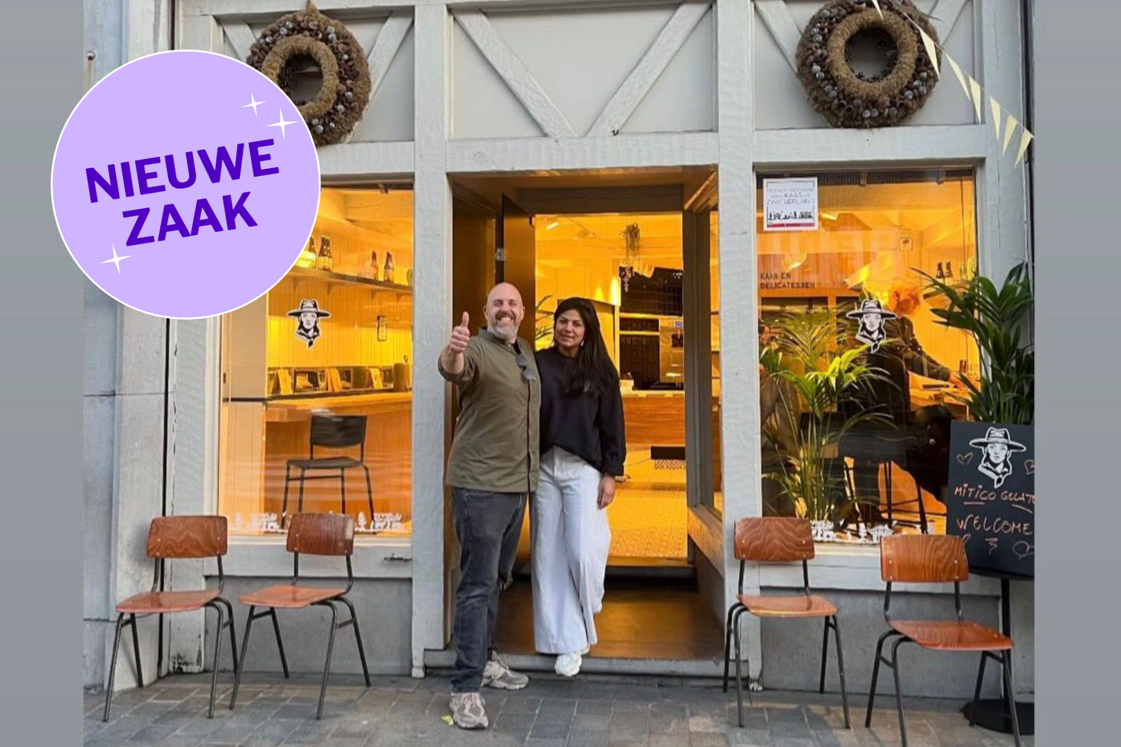 A man and woman standing at the entrance of a bakery or café, with a sign saying "Nieuwe Zaak" on the window. The man is giving a thumbs-up and smiling, while the woman stands next to him. The shop interior is visible through the window with warm lighting and plants, and there are four chairs outside.