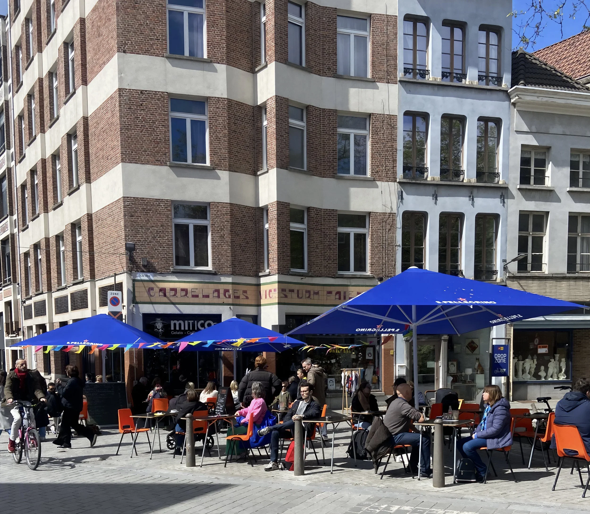 Outdoor cafe with people sitting at tables under large blue umbrellas on a city street, with buildings in the background.