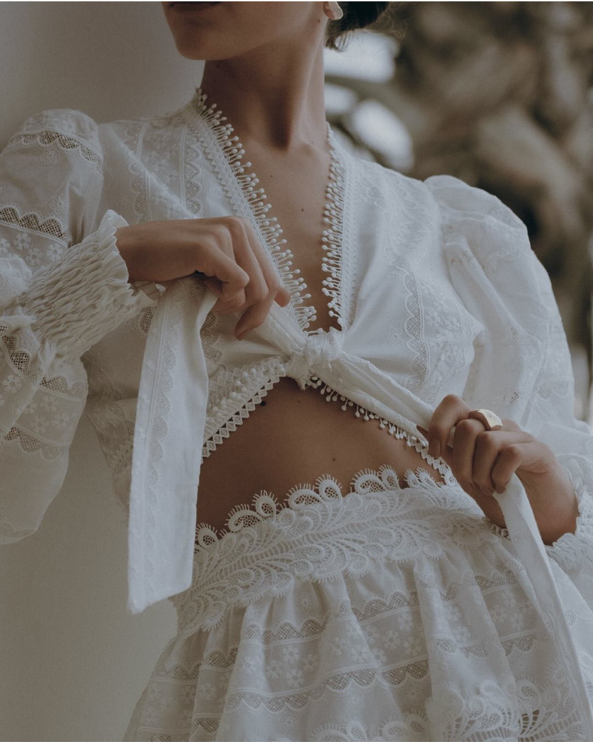 A woman in a white embroidered dress with lace details, lifting her top to reveal her midriff.