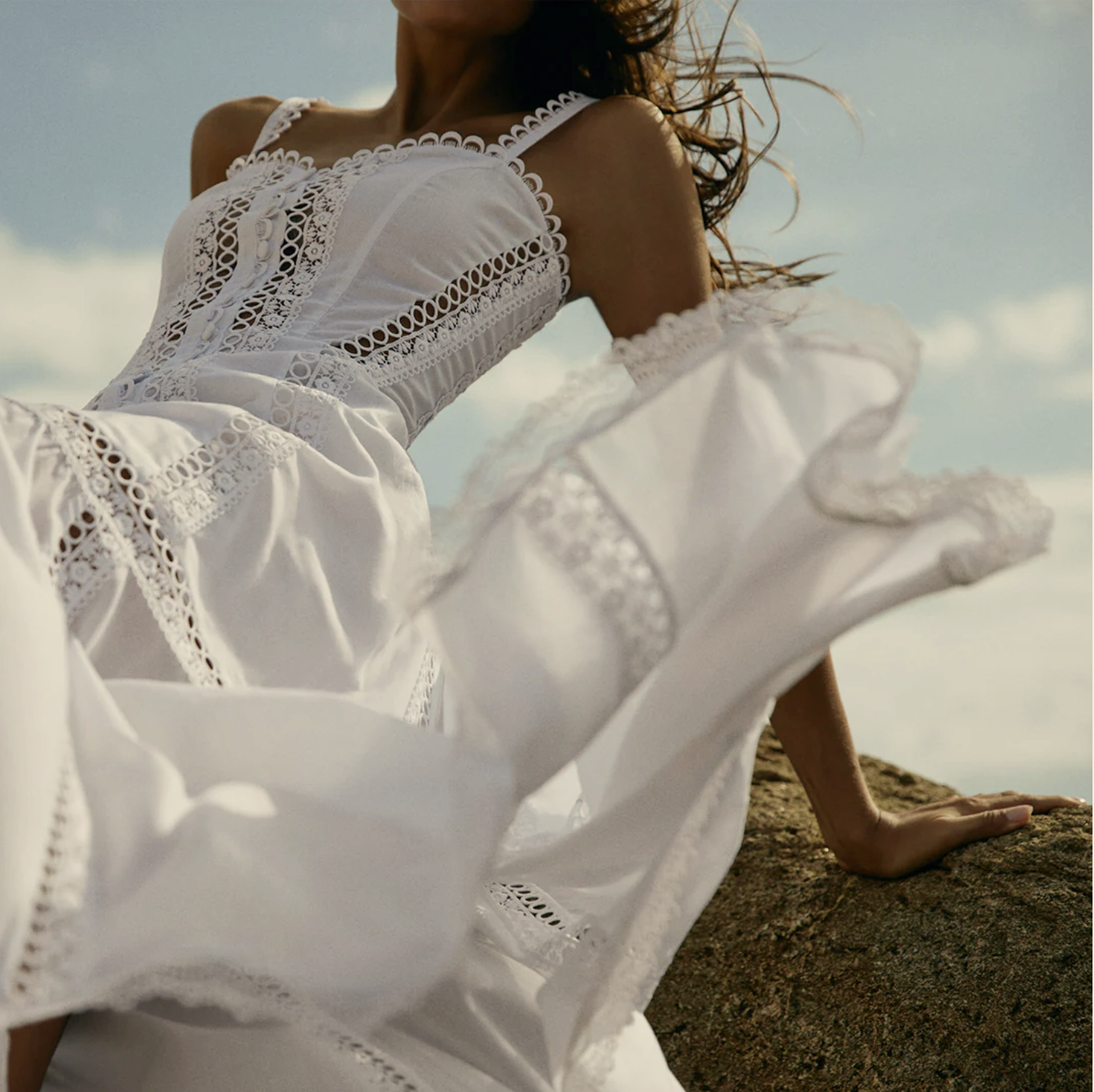 A woman in a white, embroidered sundress sitting on a rock outdoors with her hand resting on the rock and her hair blowing in the wind.