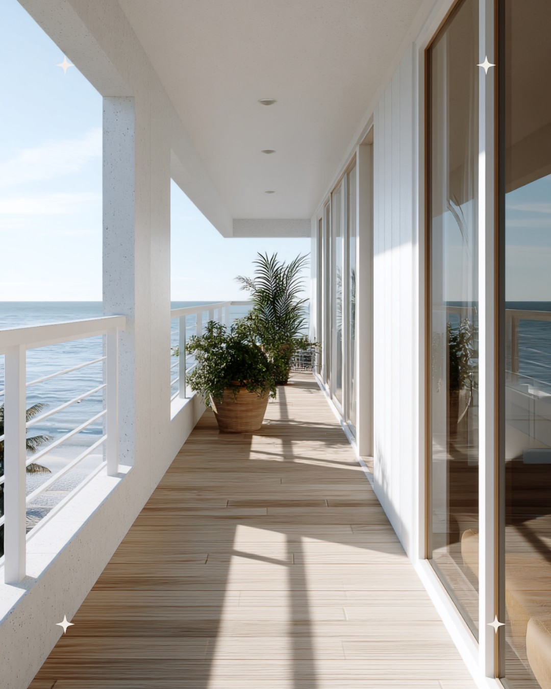 A modern beachside balcony with light wood flooring, white railings, and sliding glass doors. Potted green plants sit along the side, with ocean view in the background and blue sky overhead.