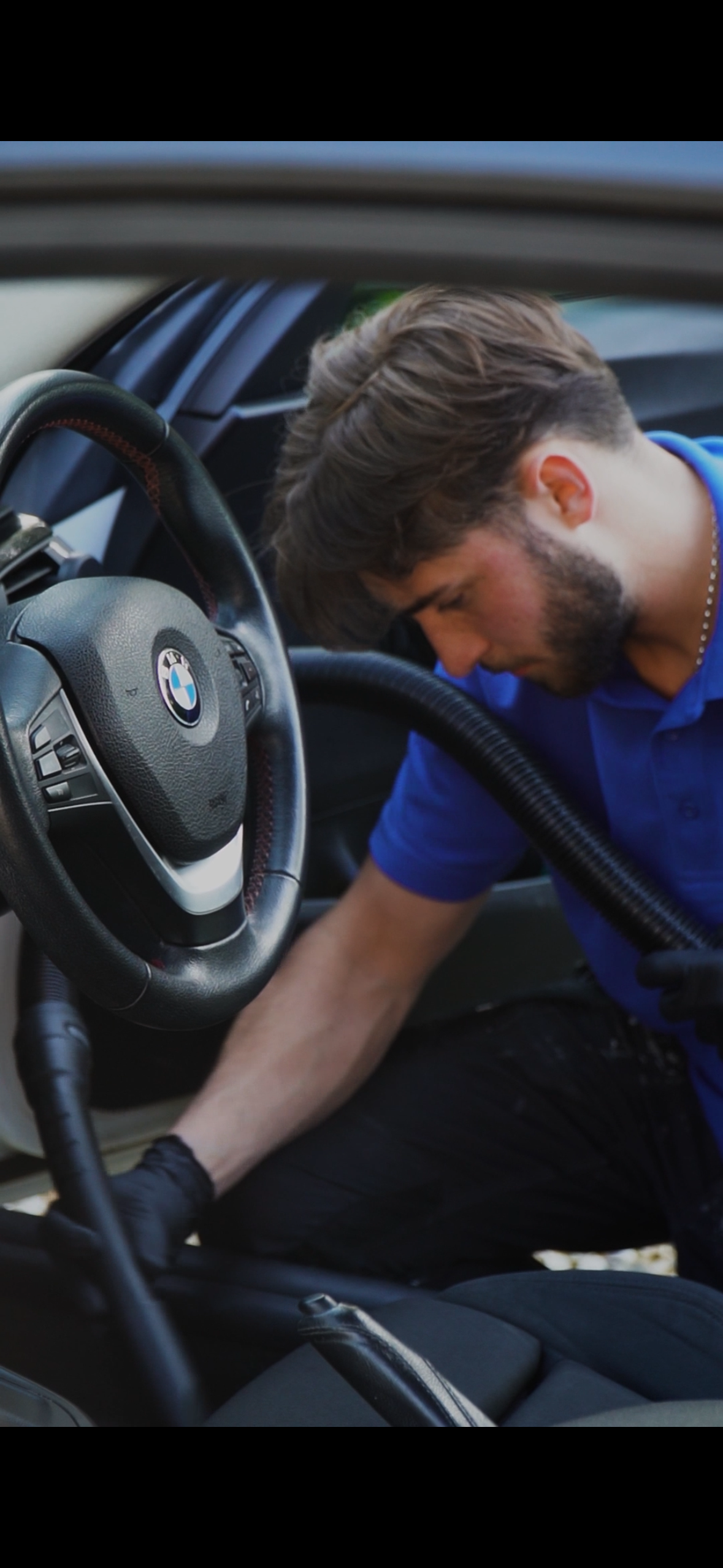 A man with a beard and brown hair, wearing a blue shirt, working inside a BMW car. He appears to be performing maintenance or repair work under the dashboard.