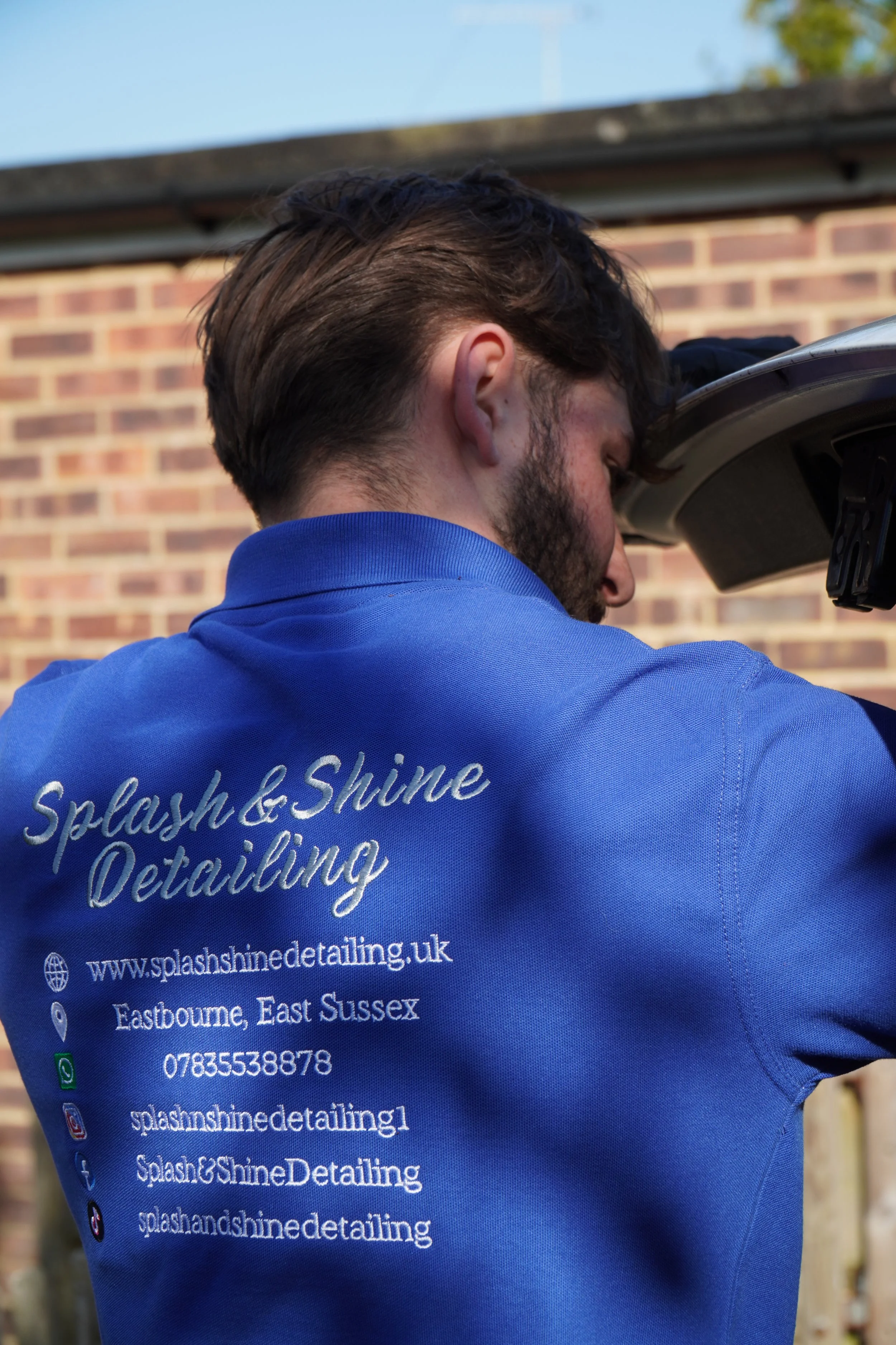 A man wearing a blue polo shirt with the text and website of Splash & Shine Detailing on the back, working on a car outside with a brick building in the background.