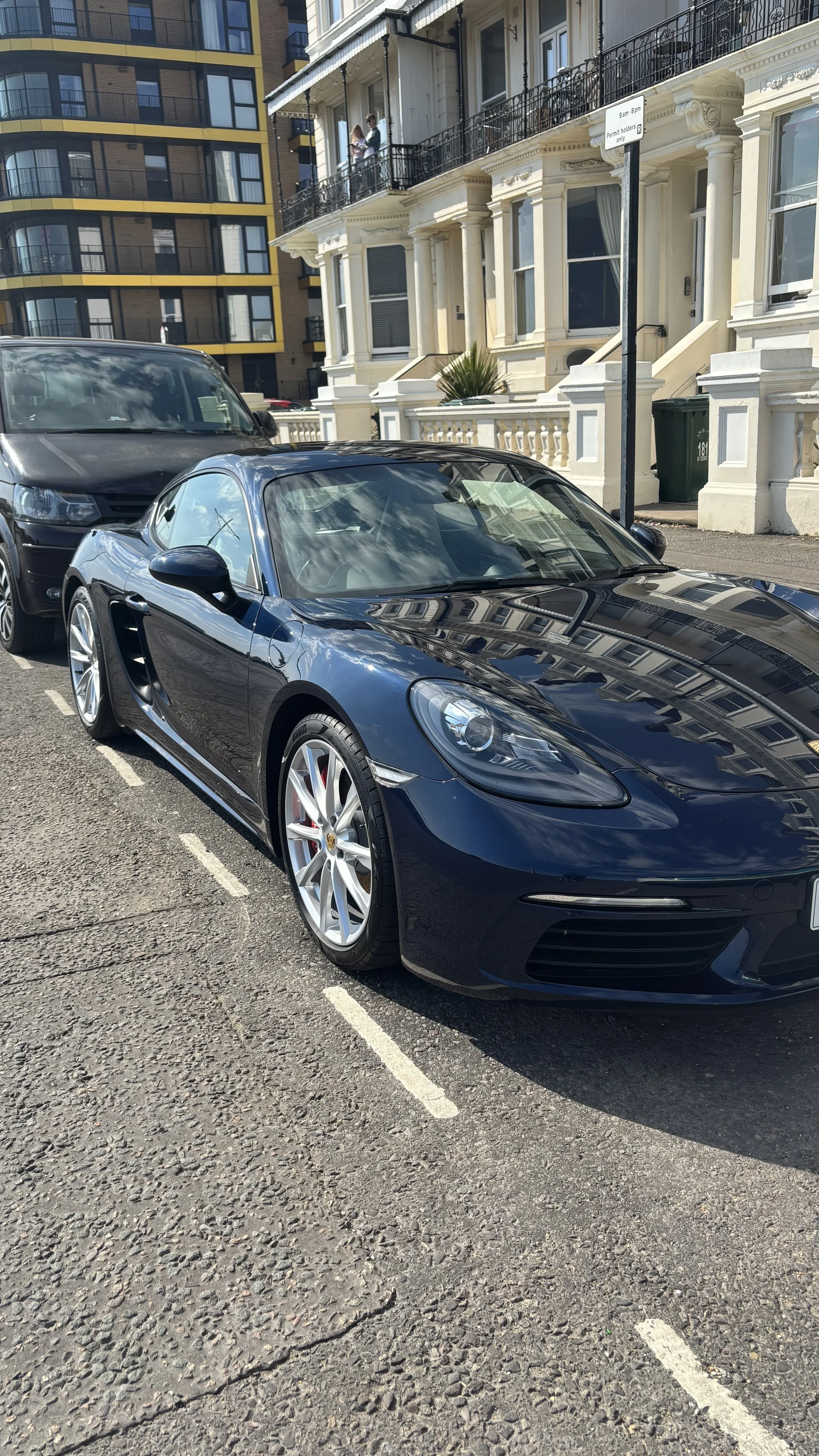 A black Porsche sports car parked on a city street in front of Victorian-style row houses, with a black van parked behind it.