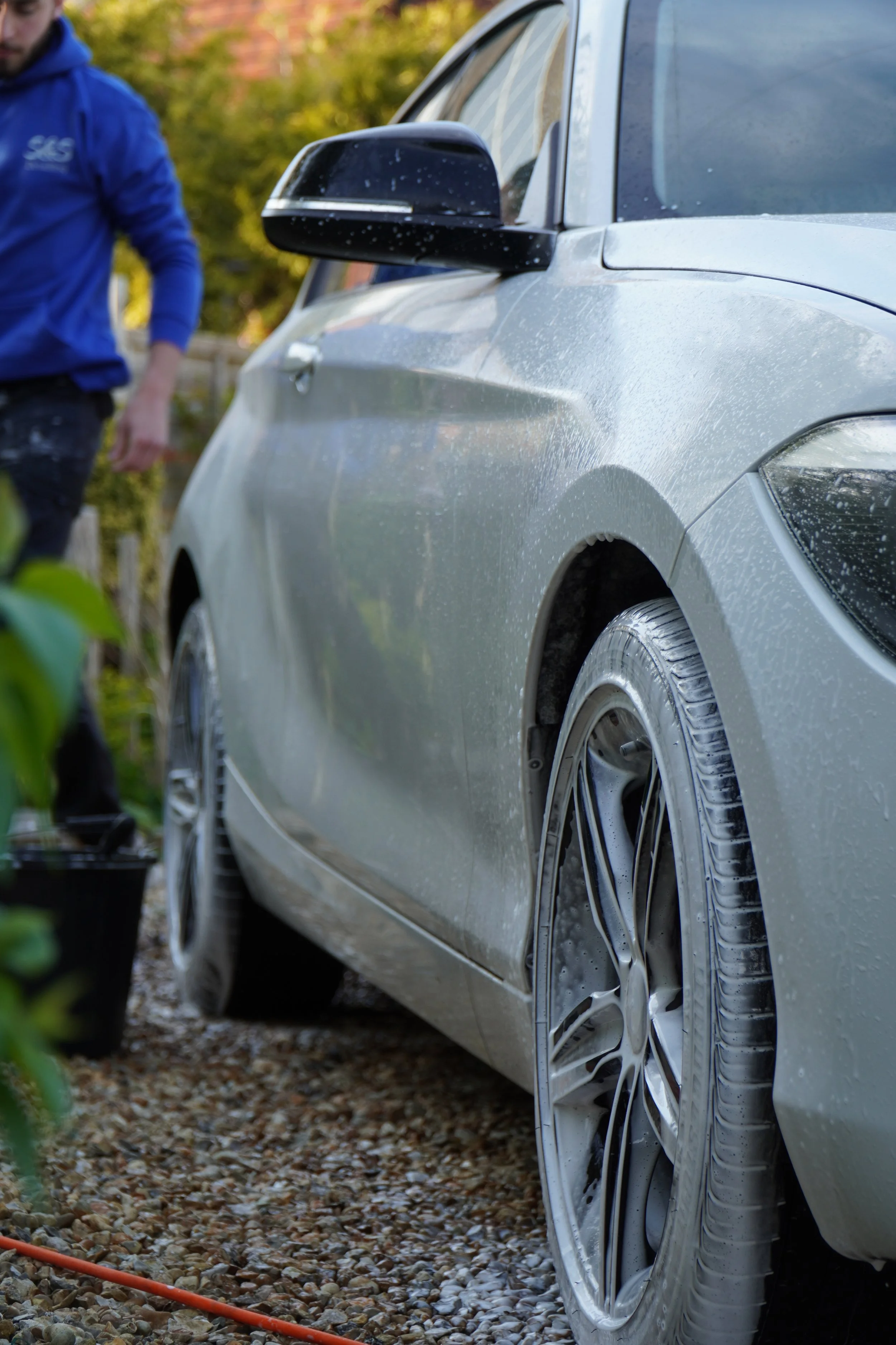 Close-up of a silver car with water droplets on its surface, parked on gravel. Part of a person in a blue jacket is visible on the left side.