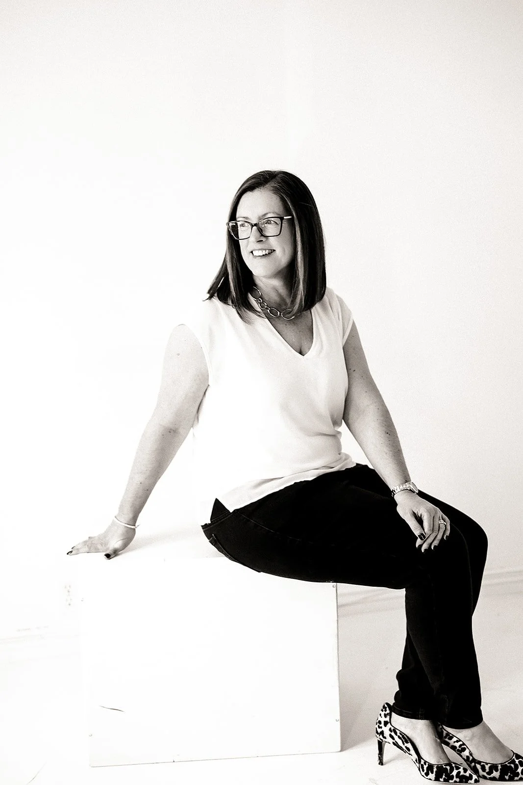A woman with glasses sitting on a white block against a plain white background, smiling and looking to her left, wearing a sleeveless top, black pants, and leopard print heels.