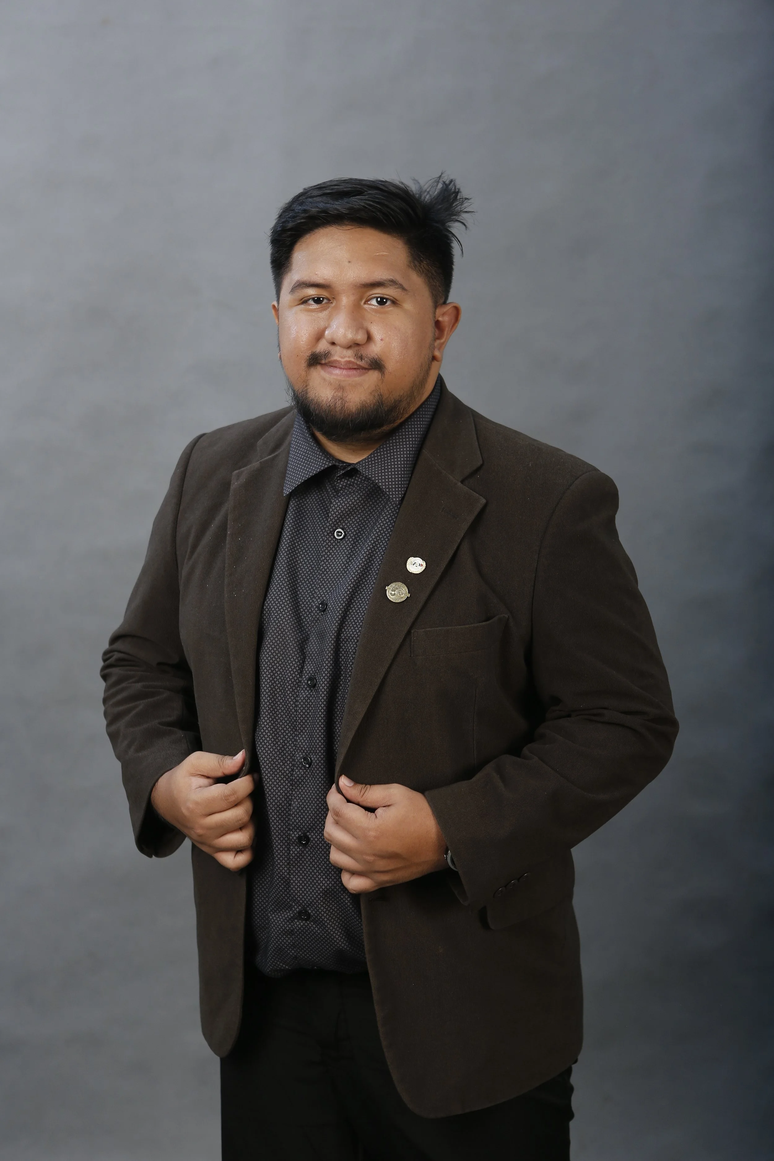 A young man with dark hair and facial hair wearing a black suit jacket, a maroon shirt, and a black tie standing against a plain beige wall.