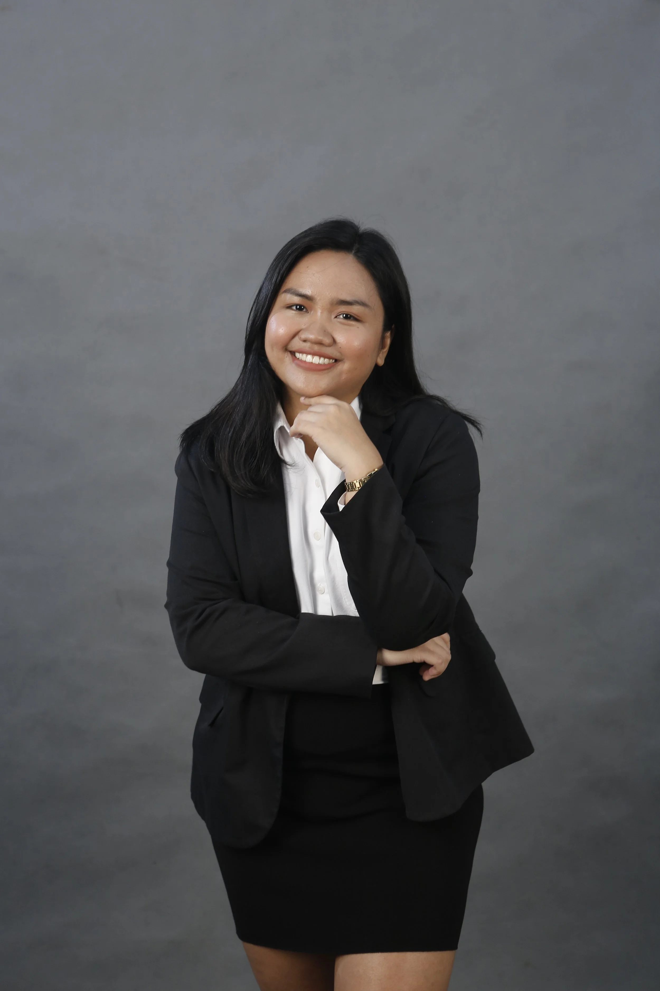 Professional headshot of a smiling young woman with long black hair, wearing a black blazer and a white top, against a white background.