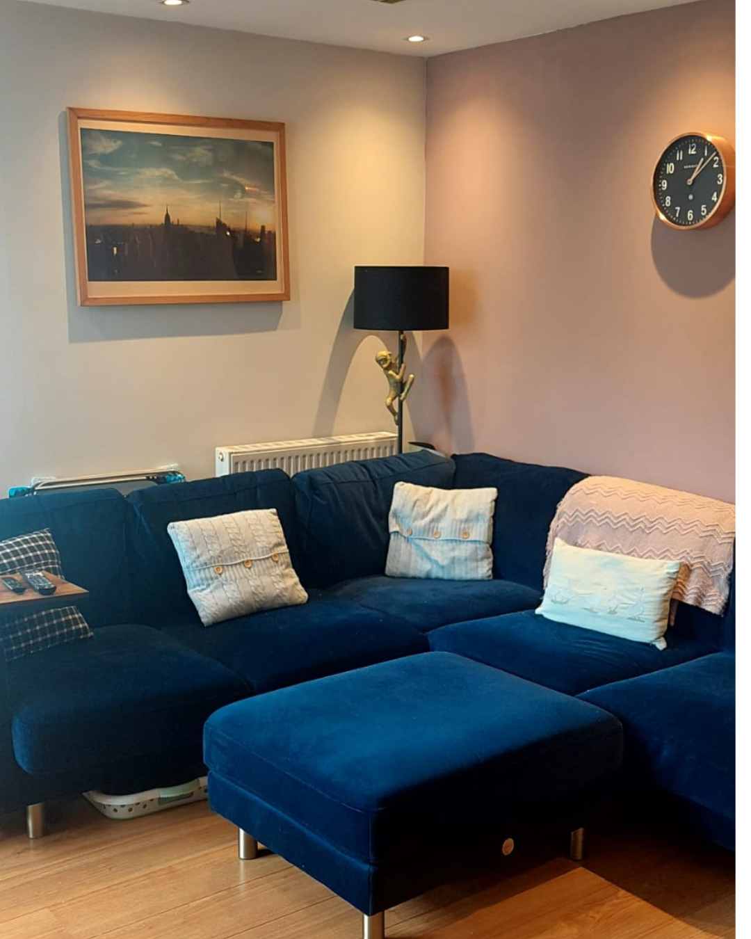 Living room with a navy blue sectional sofa, four pillows, a black floor lamp, a wall clock showing 1:09, a framed picture of a city skyline at sunset, and a wooden floor.