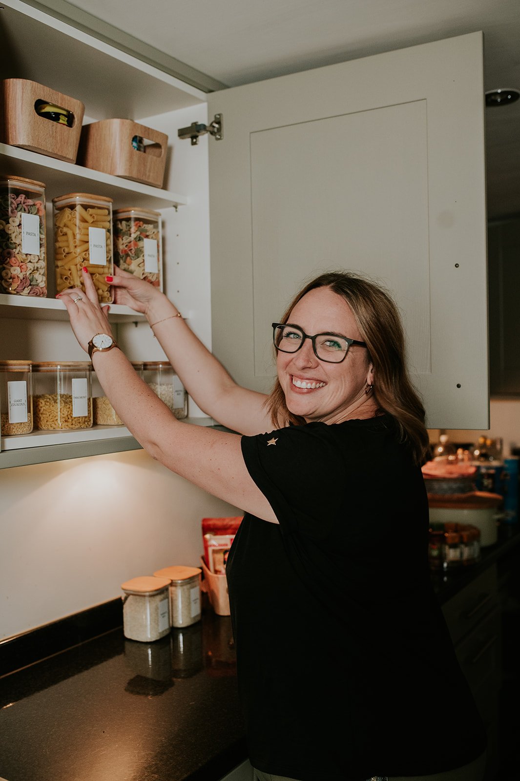 A woman with glasses and a black shirt smiling while reaching into a kitchen cabinet to grab a container of pasta. The cabinet contains various labeled containers of pasta and grains.
