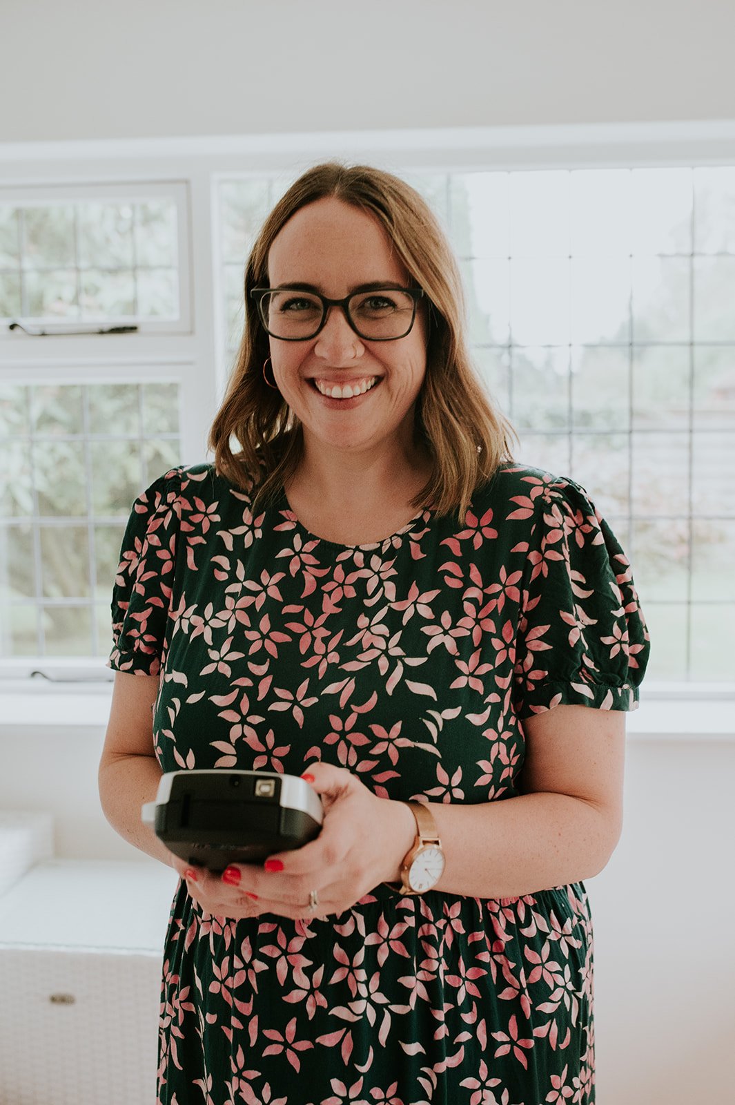 A woman with shoulder-length brown hair, glasses, and a floral dress is smiling while holding a device in her hands, standing in front of a window.