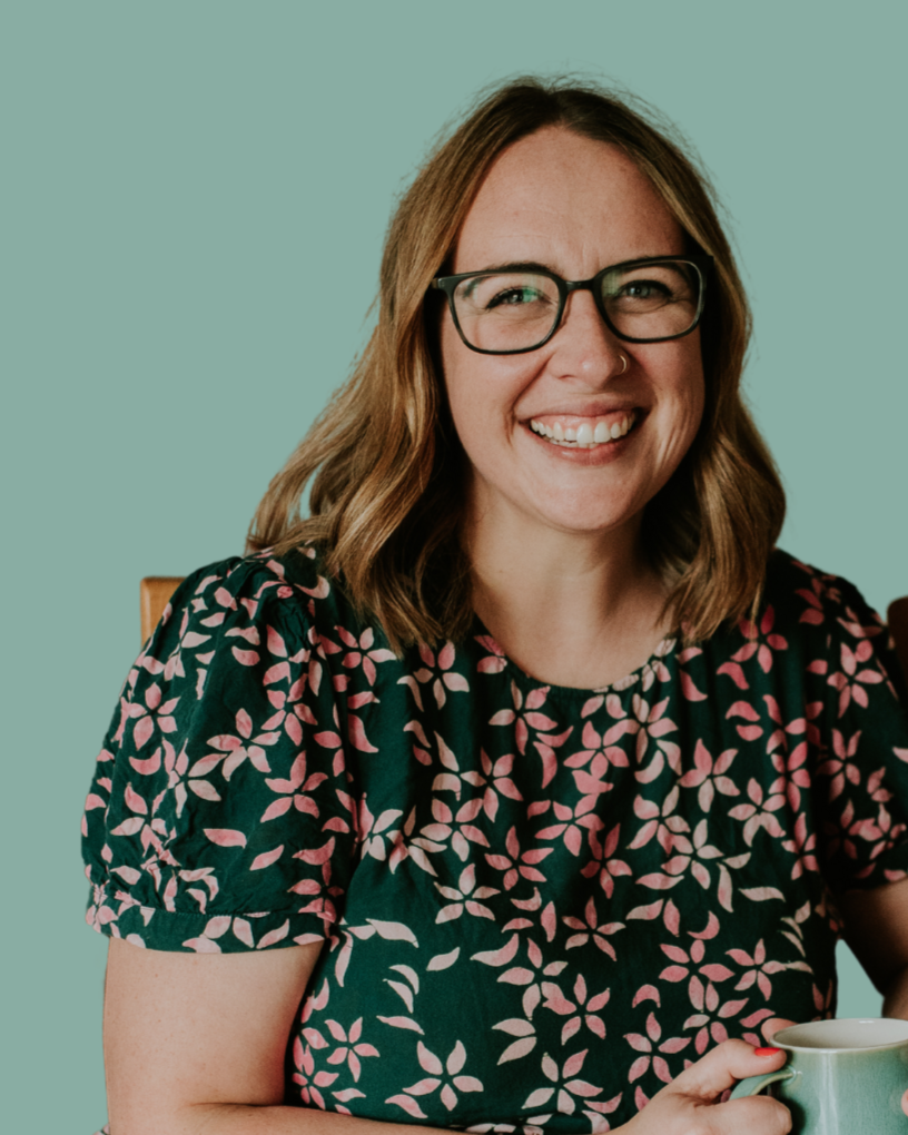 A woman with shoulder-length light brown hair, glasses, and a nose piercing, smiling and holding a mug, sitting against a plain, teal background.