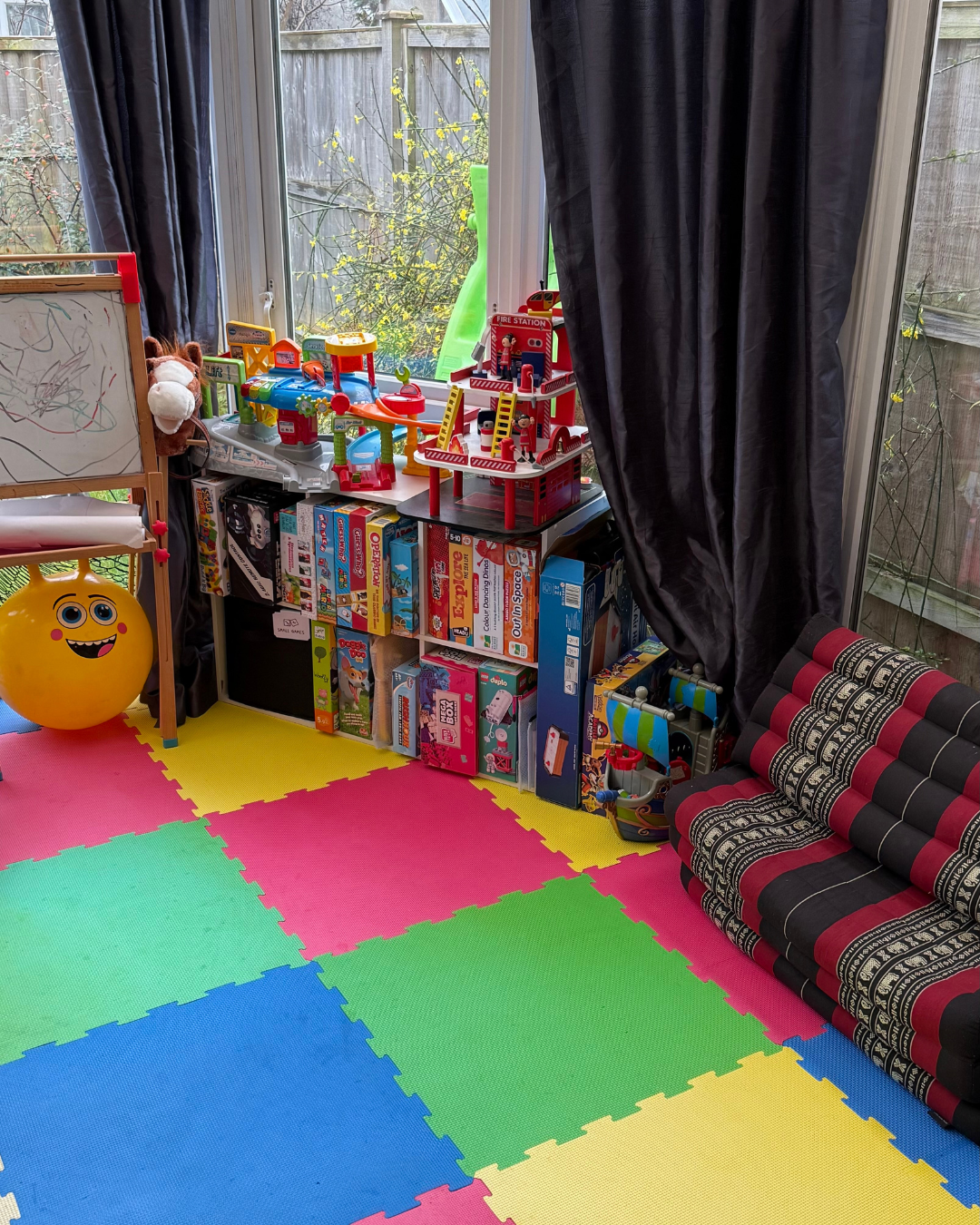 Playroom with colorful foam mats, toys including a fire station and ambulance, a yellow ball with a face, and a window with outdoor view.