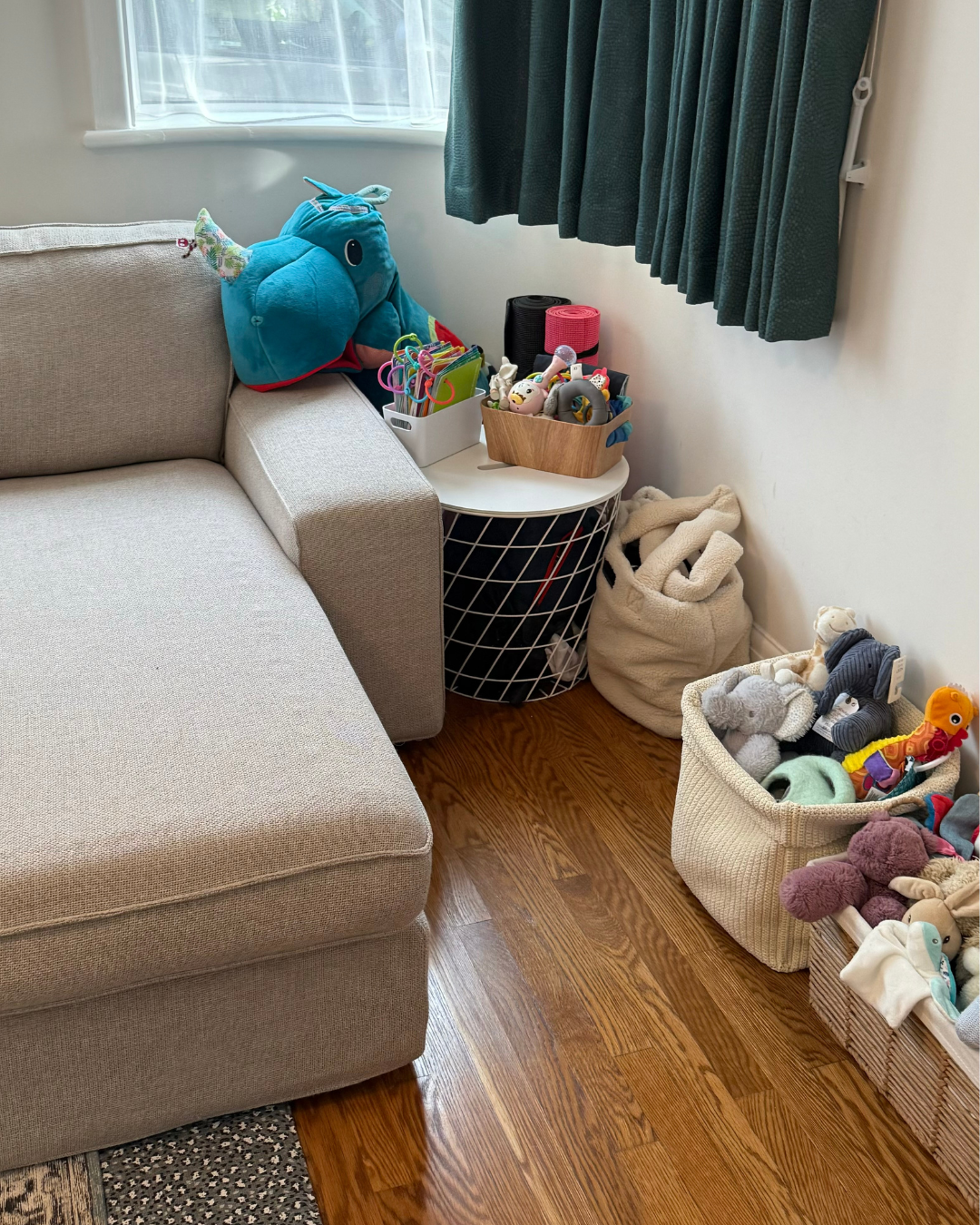 A corner of a room with a beige sectional sofa, a white side table holding toys and accessories, and baskets filled with stuffed animals and toys. There are dark green curtains and a window in the background.