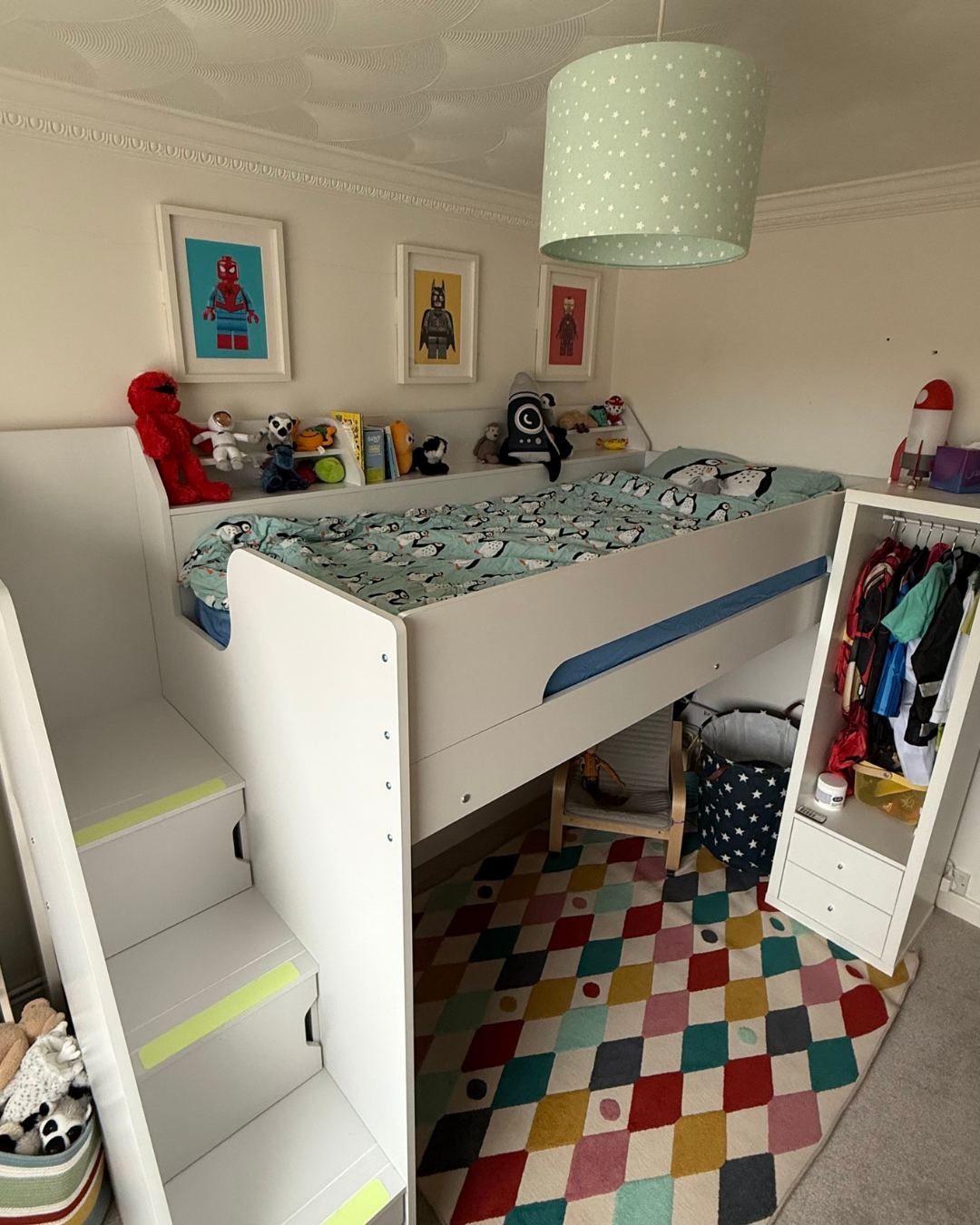 Child's bedroom with a white bed, toys, and art on the wall, including framed framed LEGO superhero prints. The bed has a penguin-patterned bedspread. There are colorful stairs, a multicolored geometric rug, and a storage unit with clothes and miscel