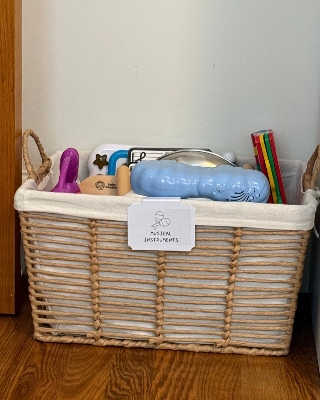 Wicker basket labeled 'Musical Instruments' contains various children's musical toys including a purple tambourine, a wooden maraca, a blue cloud-shaped instrument, a small white instrument with black star shapes, and other colorful items.