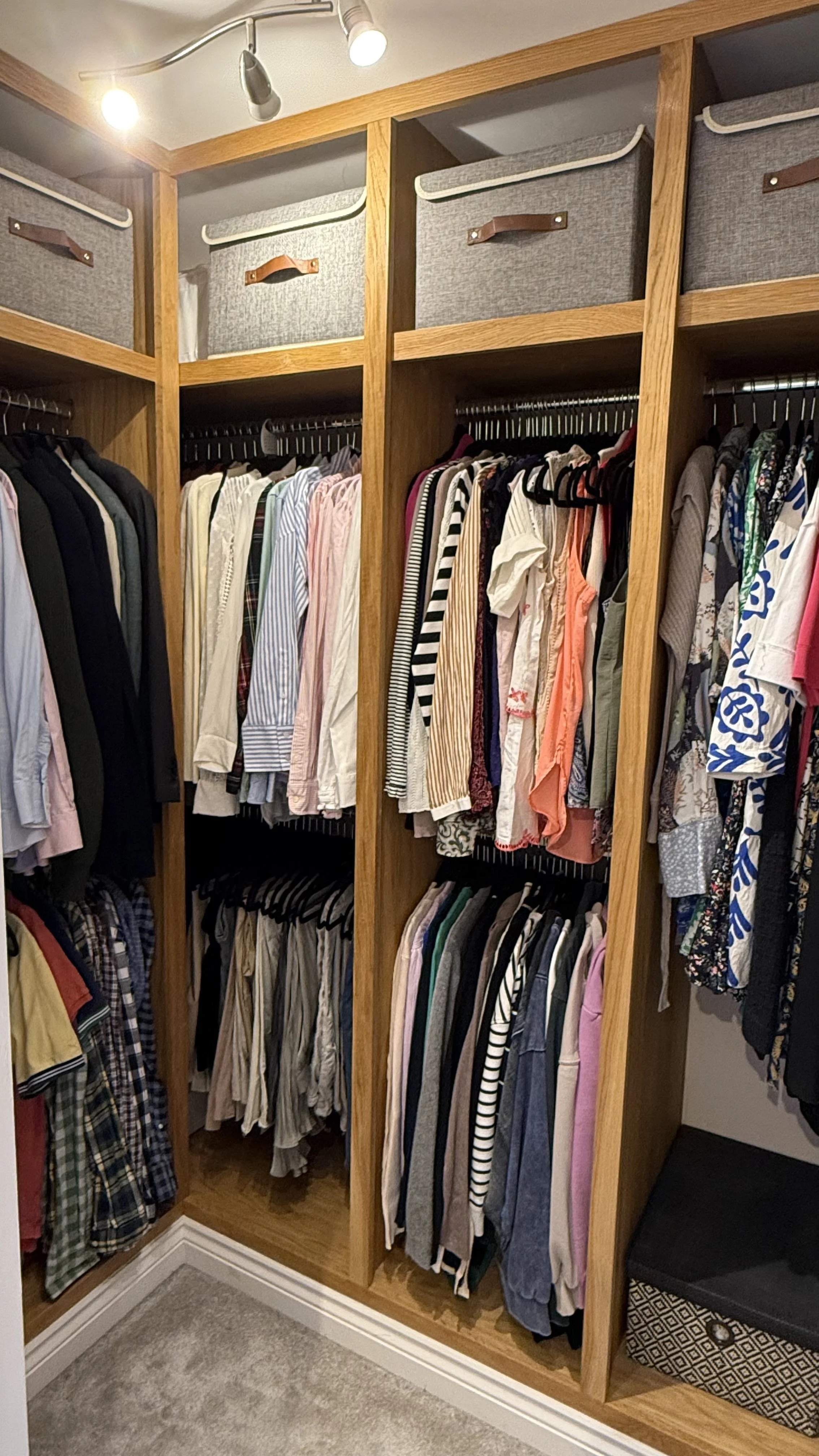 A walk-in closet with wooden shelving filled with hanging clothes and storage boxes on top.