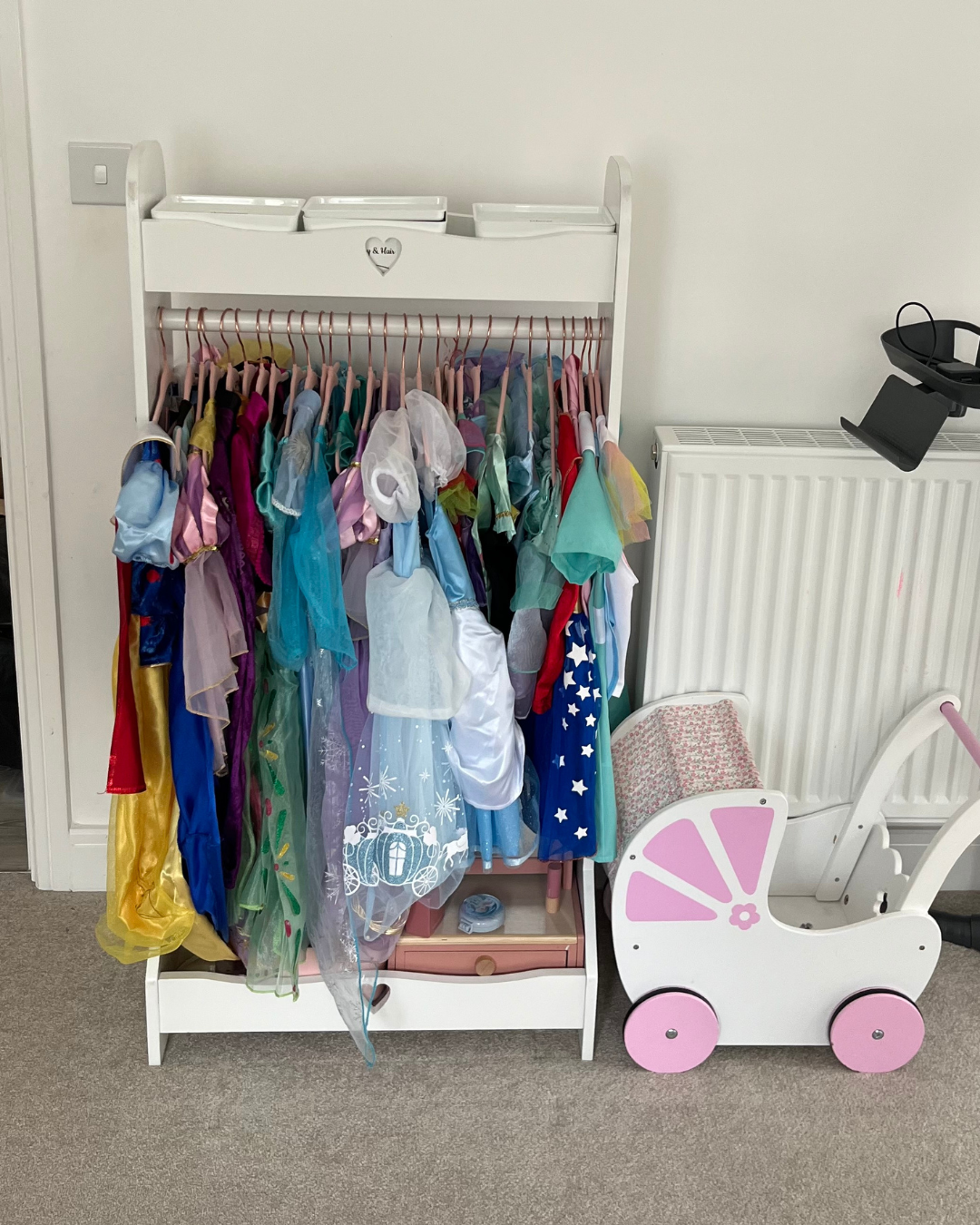 Child's dress-up corner with a white wardrobe holding colorful princess costumes and a pink toy stroller next to a white radiator.