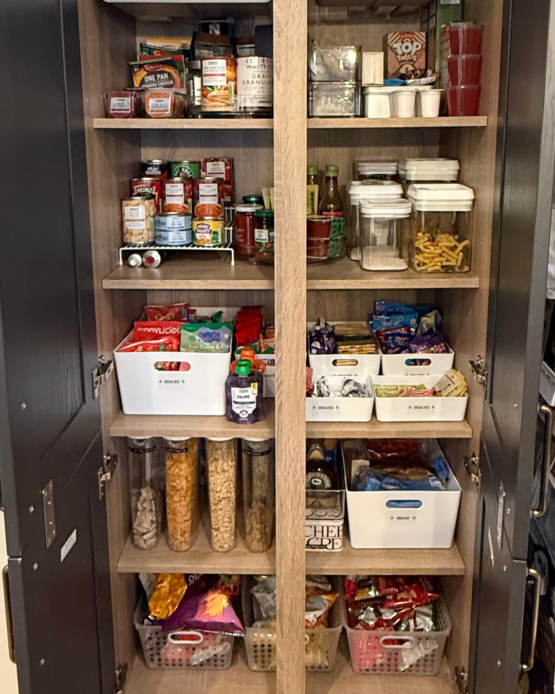 Pantry cabinet filled with snacks, canned goods, condiments, cereal, and packaged food stored on wooden shelves.