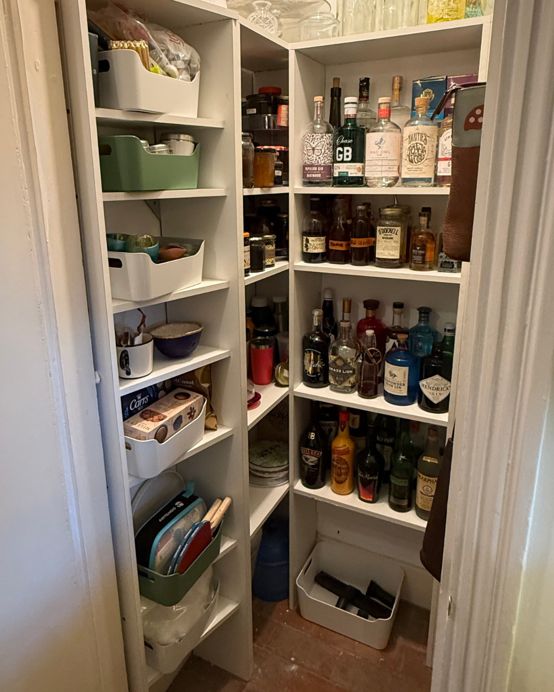 A narrow pantry closet with shelves holding various bottles of alcohol, jars, glasses, snacks, and kitchen items.
