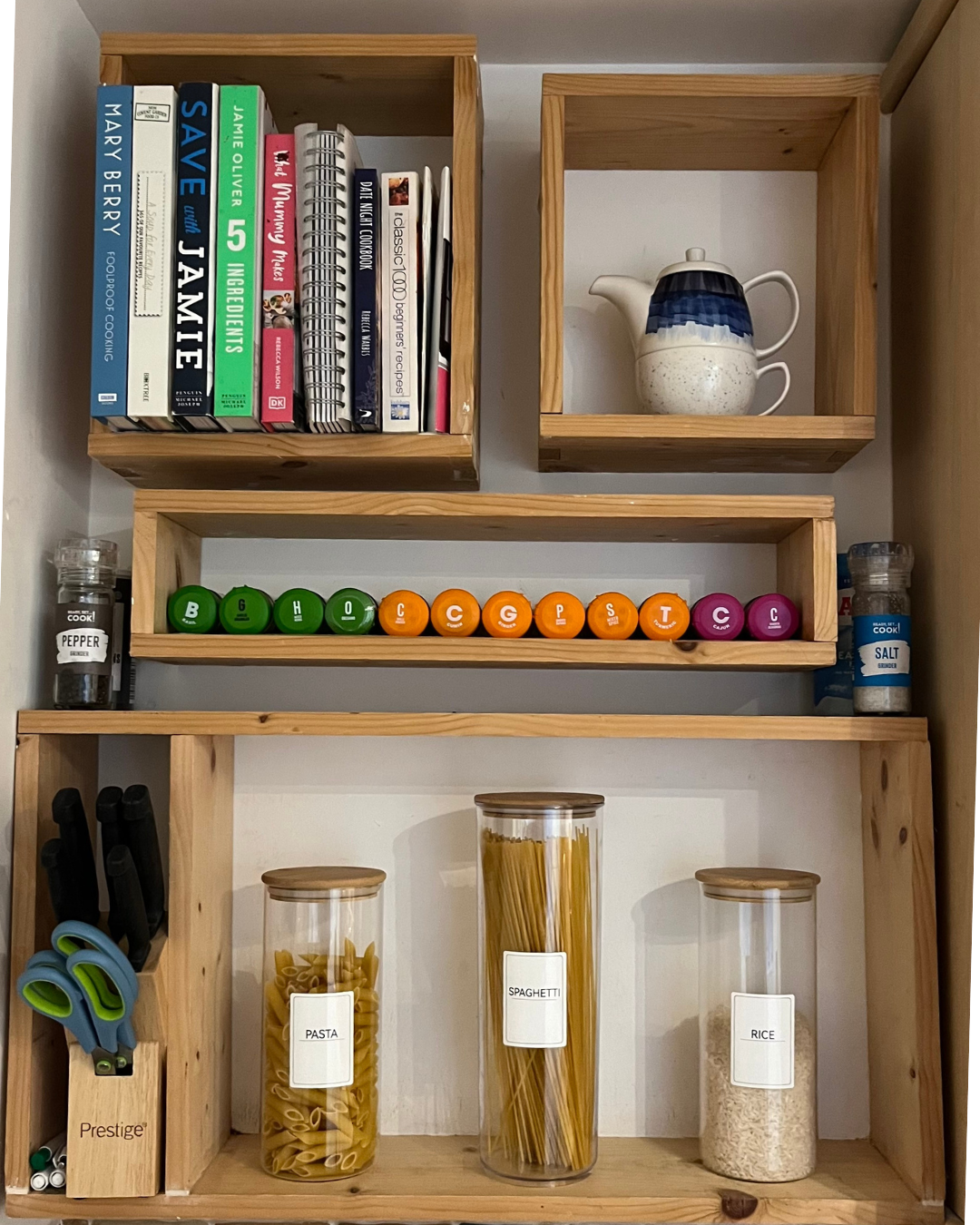 Wooden shelves holding cookbooks, a ceramic teapot, colorful containers, a salt shaker, a pepper grinder, scissors, pens, and jars of pasta, spaghetti, and rice in a kitchen.