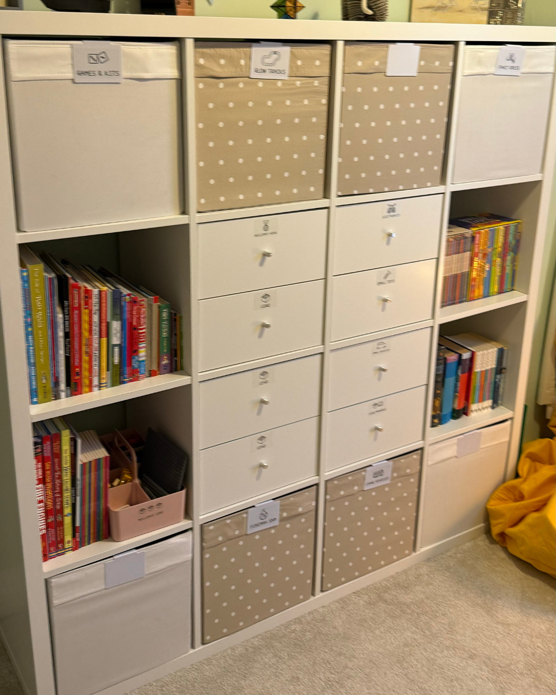 A white bookshelf with labeled storage bins, books, and a pink storage box, positioned against a wall with a green wall nearby.