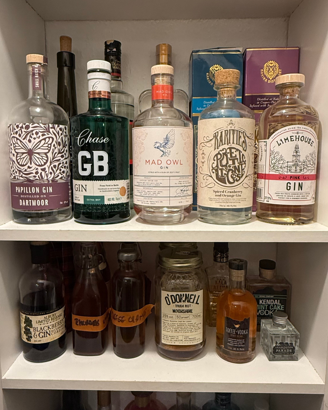 A collection of various bottles of gin, whiskey, and vodka on a white shelf, including brands like Papillon Gin, Chase GB Gin, Mad Owl Gin, and Limehouse Gin.