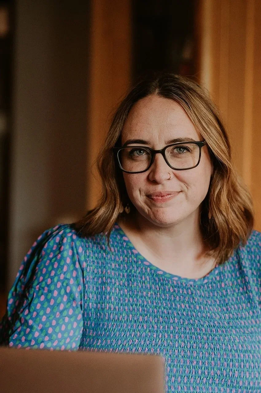 A woman with shoulder-length brown hair, wearing glasses, a nose ring, and a colorful blue, purple, and green patterned top, smiling in front of a wooden background.
