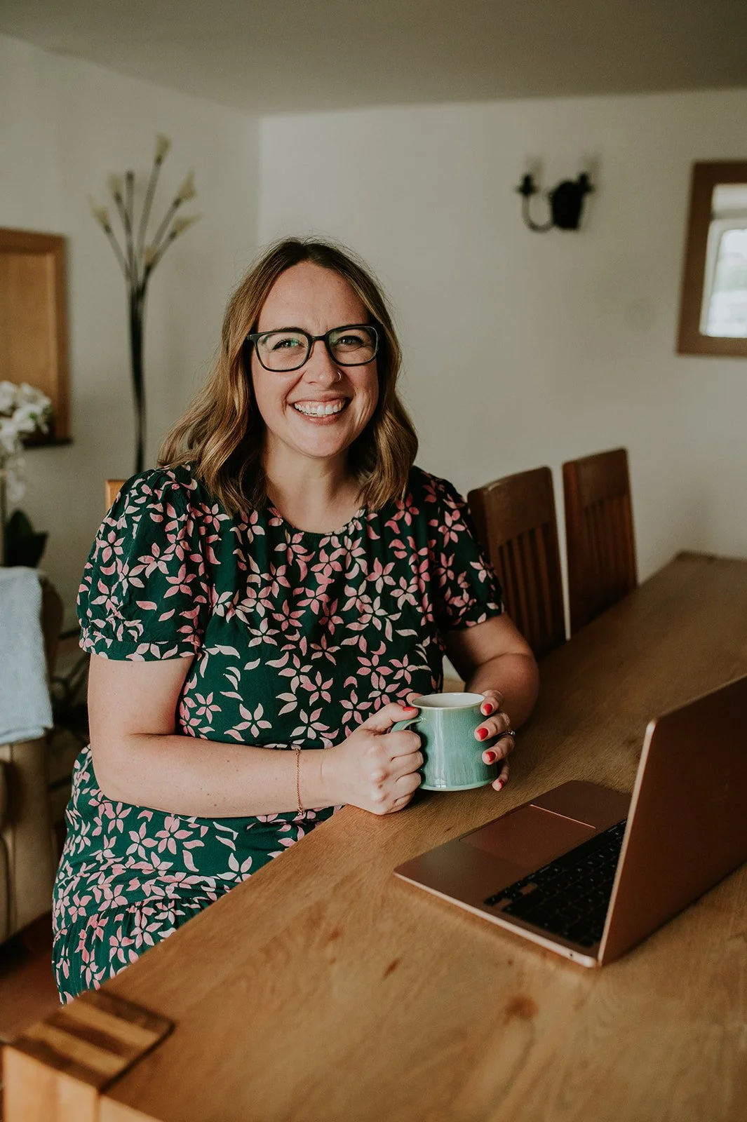 A woman with shoulder-length brown hair, glasses, and a floral dress sitting at a wooden table with a laptop and holding a mug.