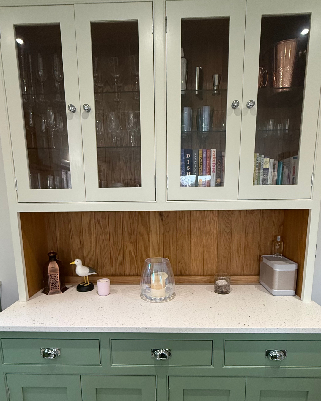 A vintage-style green and white china cabinet with glass doors, displaying glassware and cookbooks. The lower section has a white countertop with decorative items including a lantern, a seagull figure, a candle, a glass lantern with a candle, a small