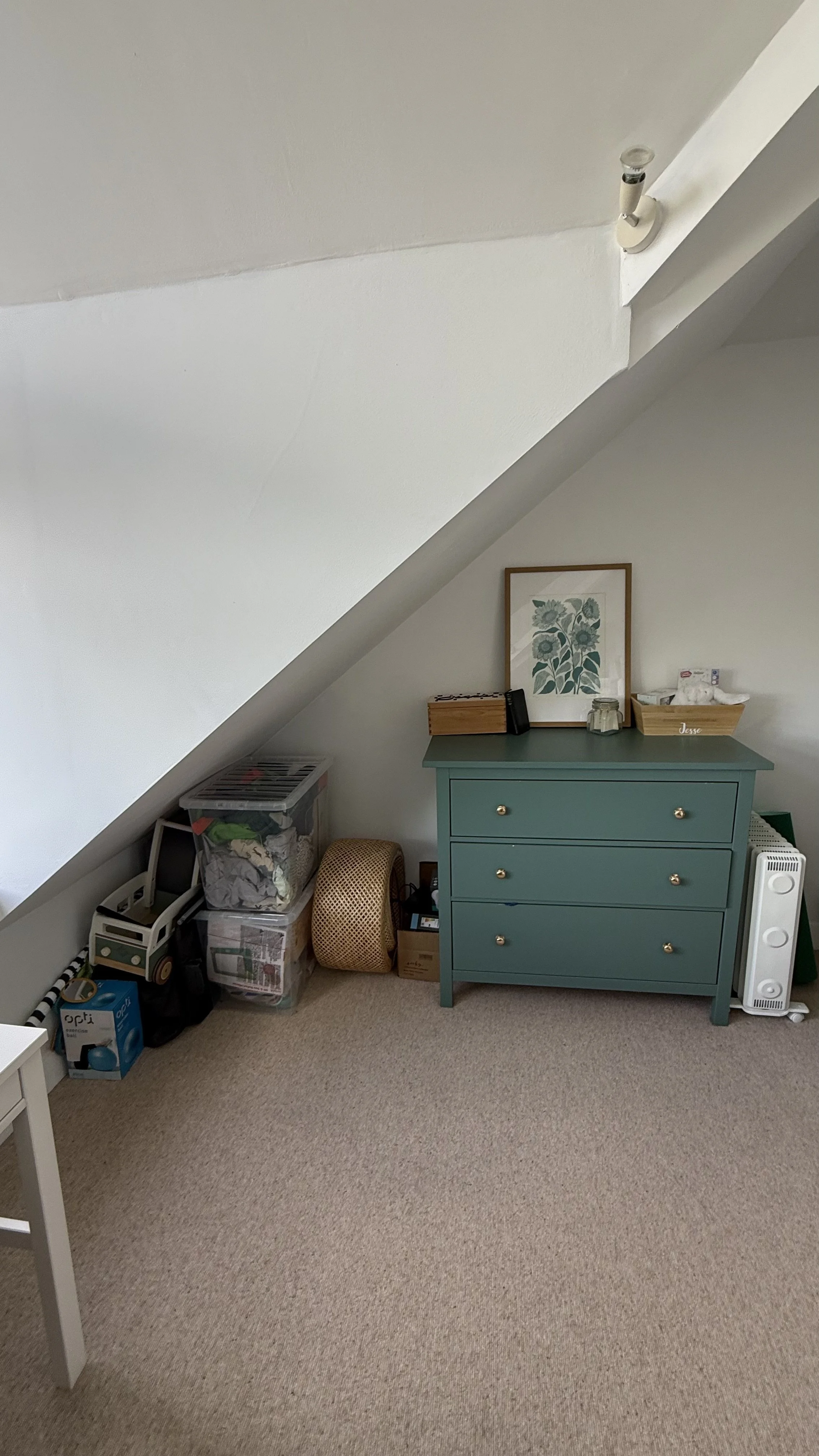 A corner of a room with sloped ceiling, a teal dresser with three drawers, a framed botanical print on top, and various storage boxes and items to the left of the dresser. A small white chair is partially visible in the foreground.