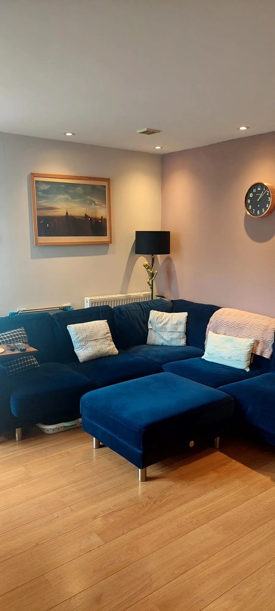 A living room with a blue sectional sofa, decorative pillows, a framed landscape photograph on the wall, a black floor lamp with a gold sculpture base, and a wall clock. The floor is wood, and there's a radiator under the photograph.