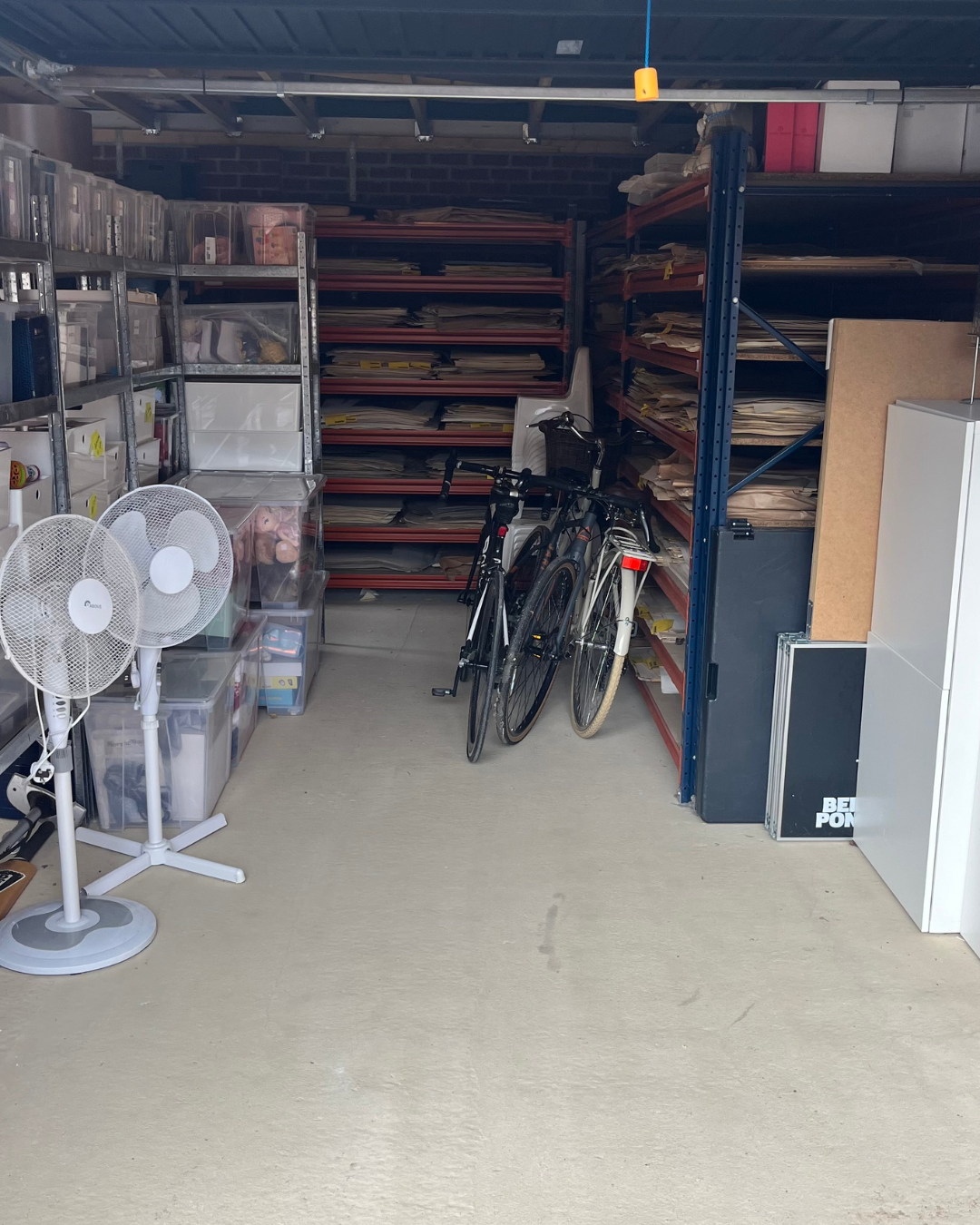 A storage room with metal shelves filled with files and folders, two bicycles parked, two white oscillating fans, plastic bins with miscellaneous items, a white cabinet, and other storage boxes.