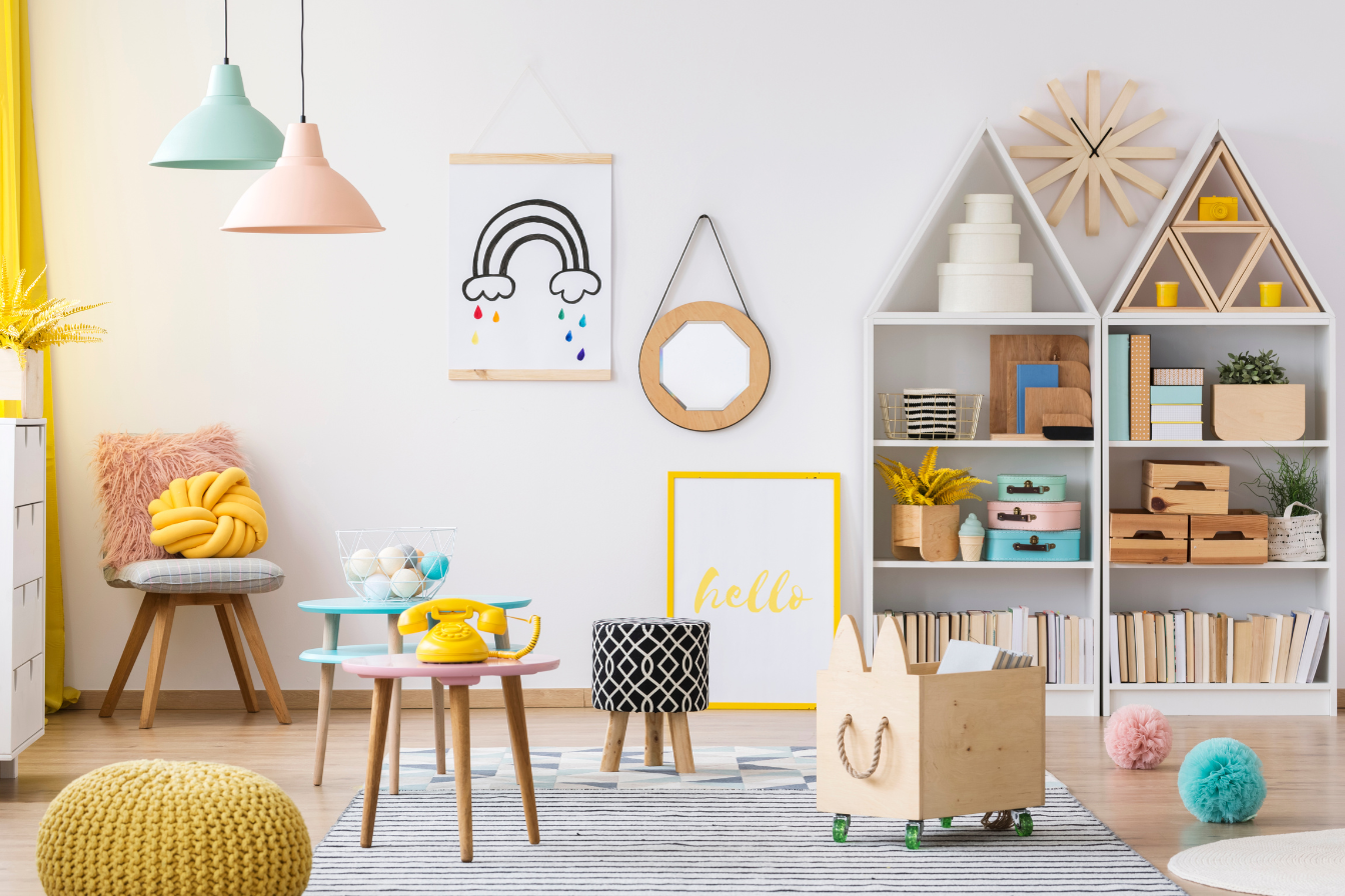 Colorful, modern children's playroom with pastel-colored pendant lights, decorative wall art, bookshelves with storage, a woven pouf, a wooden rolling cart, and playful soft balls on the floor.