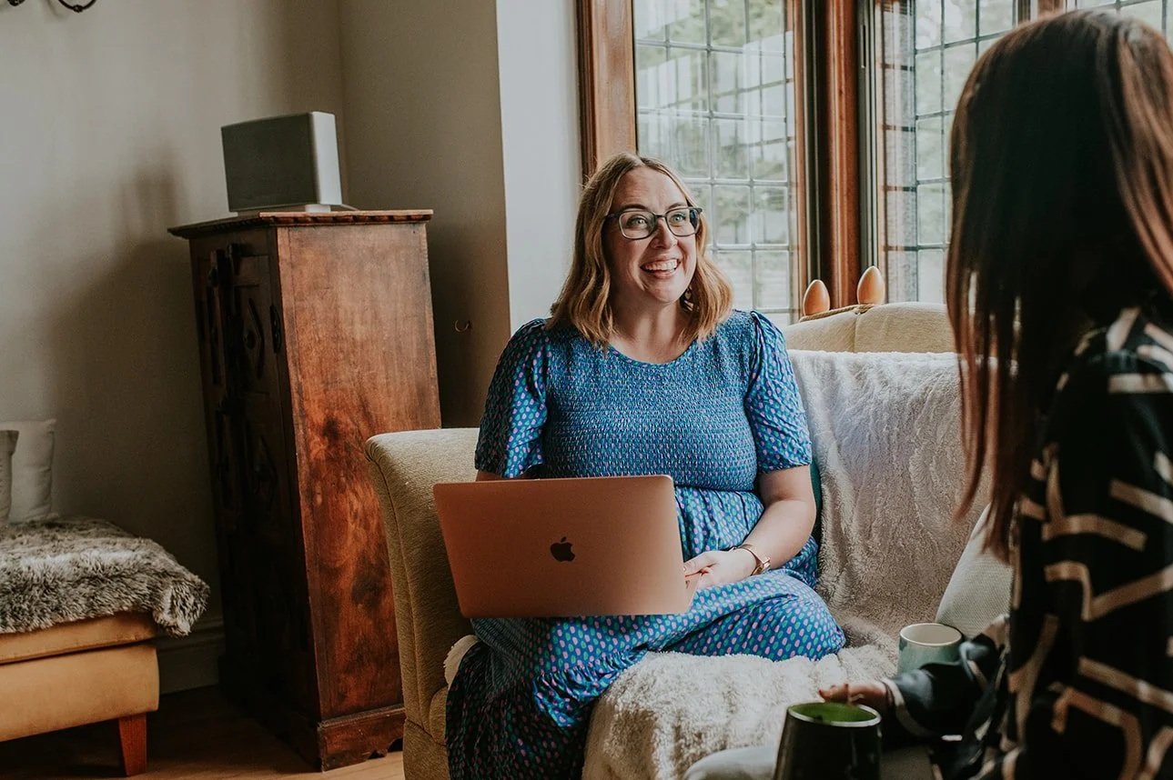 A woman with shoulder-length blonde hair and glasses smiling while sitting on a beige couch with a laptop on her lap, talking to another woman with long reddish-brown hair. They are in a cozy room with large windows and natural light.